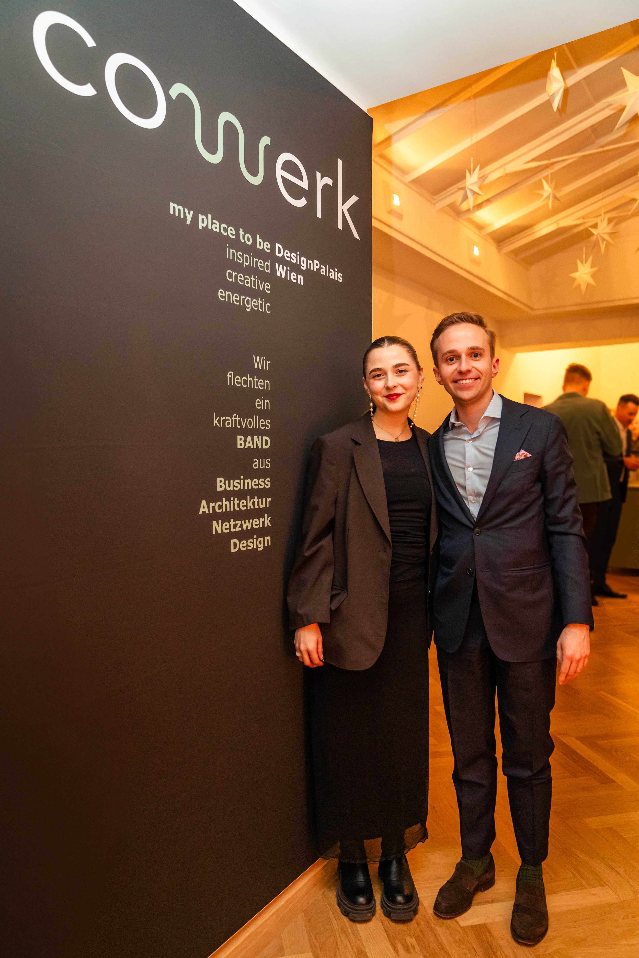 Zwei Personen, eine Frau und ein Mann, stehen vor einem schwarzen Wandbild mit Text über DesignPalais in Wien, lächeln für die Kamera und tragen formelle Kleidung.