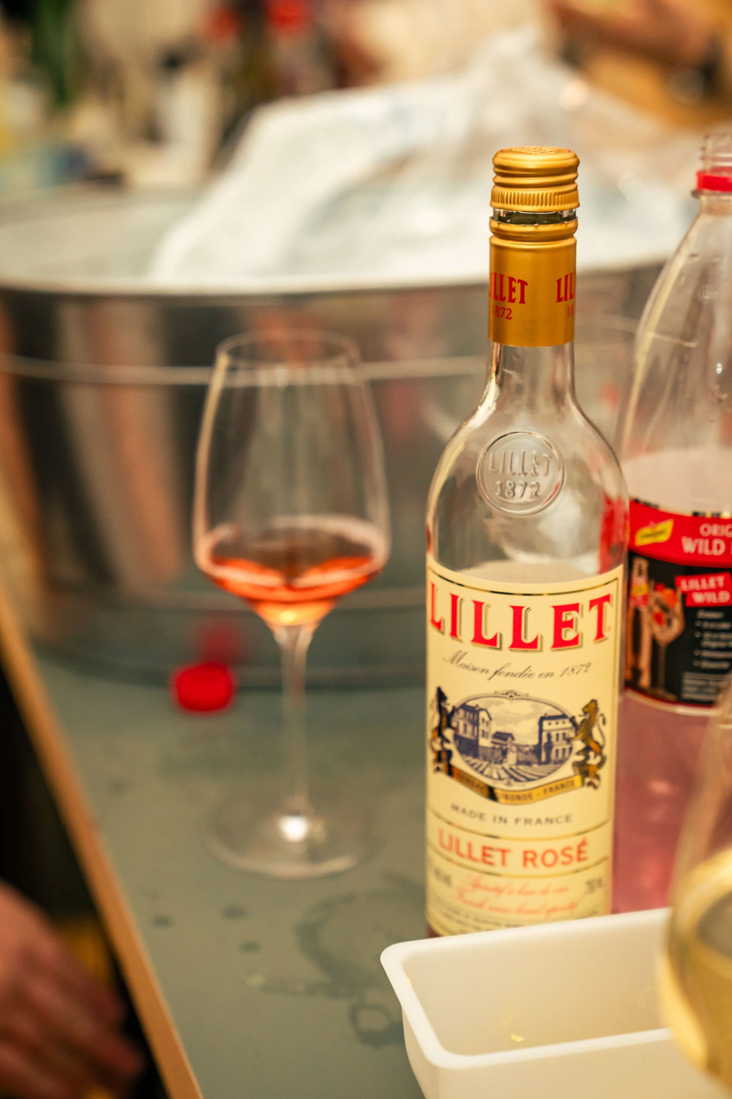 Eine Flasche Lillet Rosé und ein Weinglas mit rosafarbenem Getränk stehen auf einem Tisch mit einem großen Metallbehälter im Hintergrund.