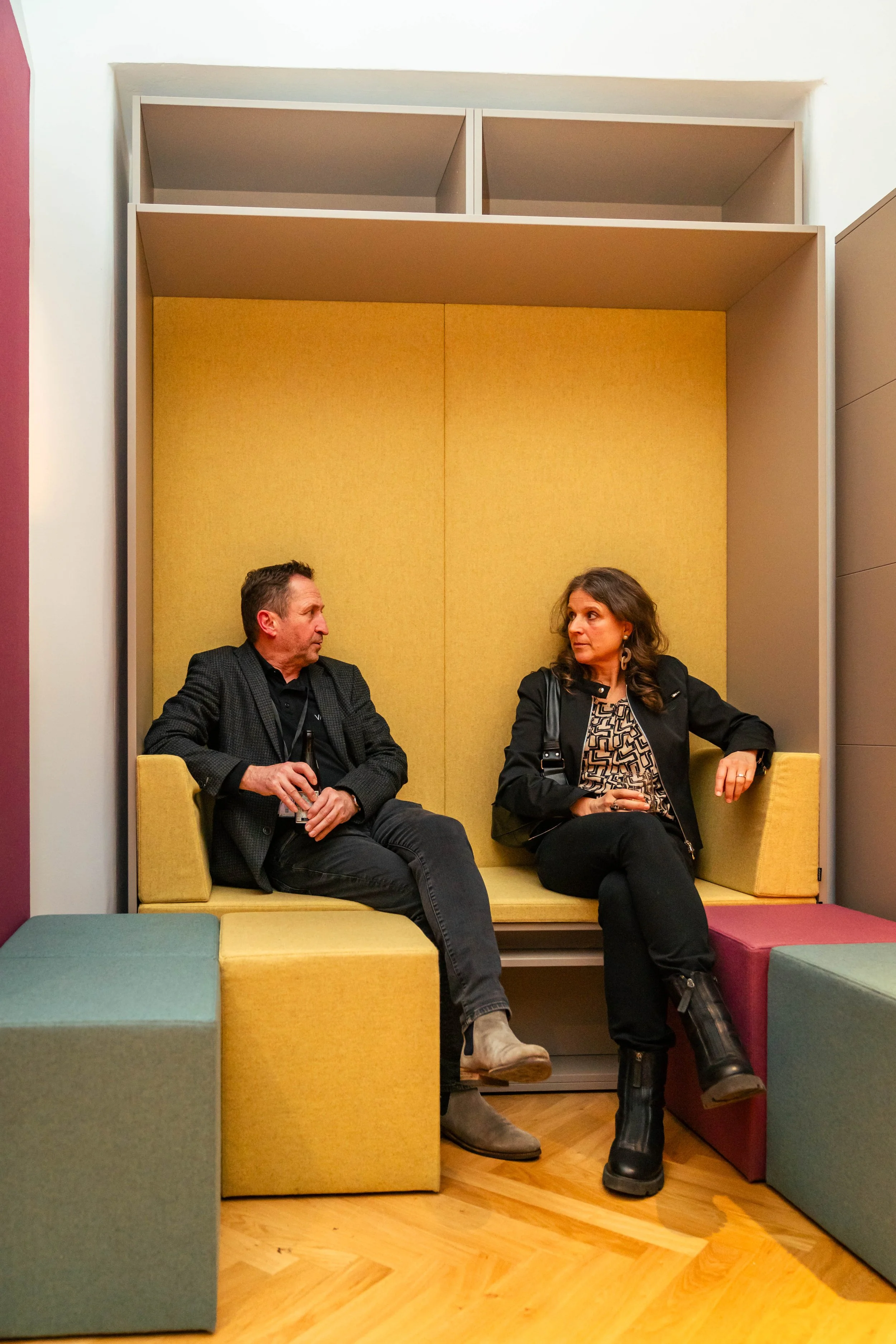Zwei Menschen sitzen in einem Loungebereich, die links ein Mann mit dunkler Jacke und grauen Stiefeln, rechts eine Frau mit schwarzer Jacke und schwarzen Stiefeln, im Hintergrund eine gelbe Wand und bunte Sitzwürfel.