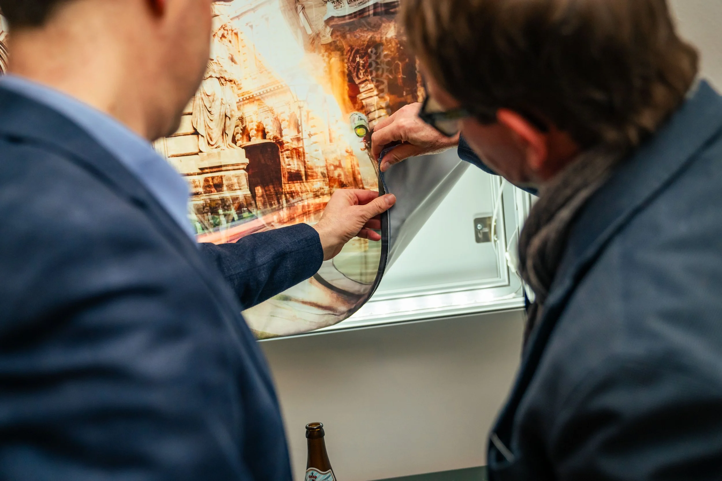 Zwei Männer betrachten eine große fotografische Poster, das den Eingang des Kolosseums in Rom zeigt, in einem Raum mit Fenster. Einer der Männer zieht eine Ecke des Posters zurück, während der andere zusieht.