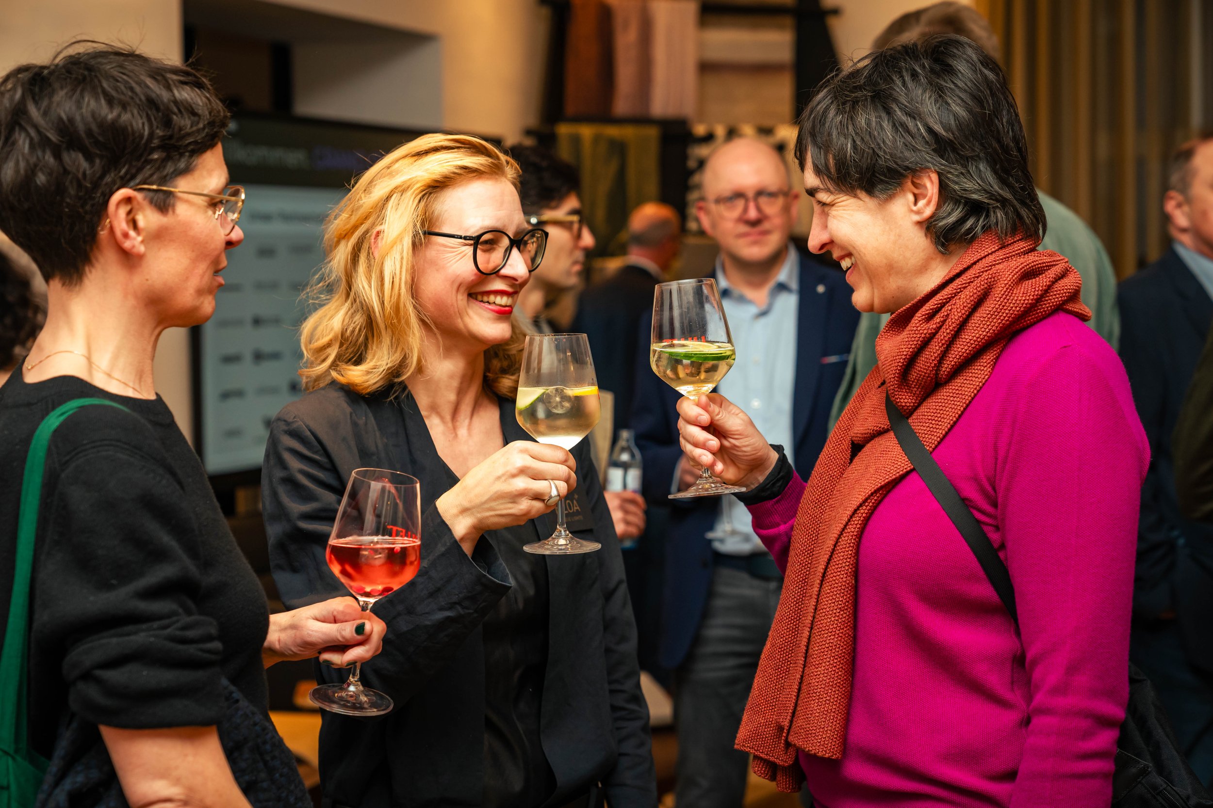 Drei Frauen beim Anstoßen mit Gläsern Wein auf einer gesellschaftlichen Veranstaltung, miteinander lachend und in gepflegter Kleidung.