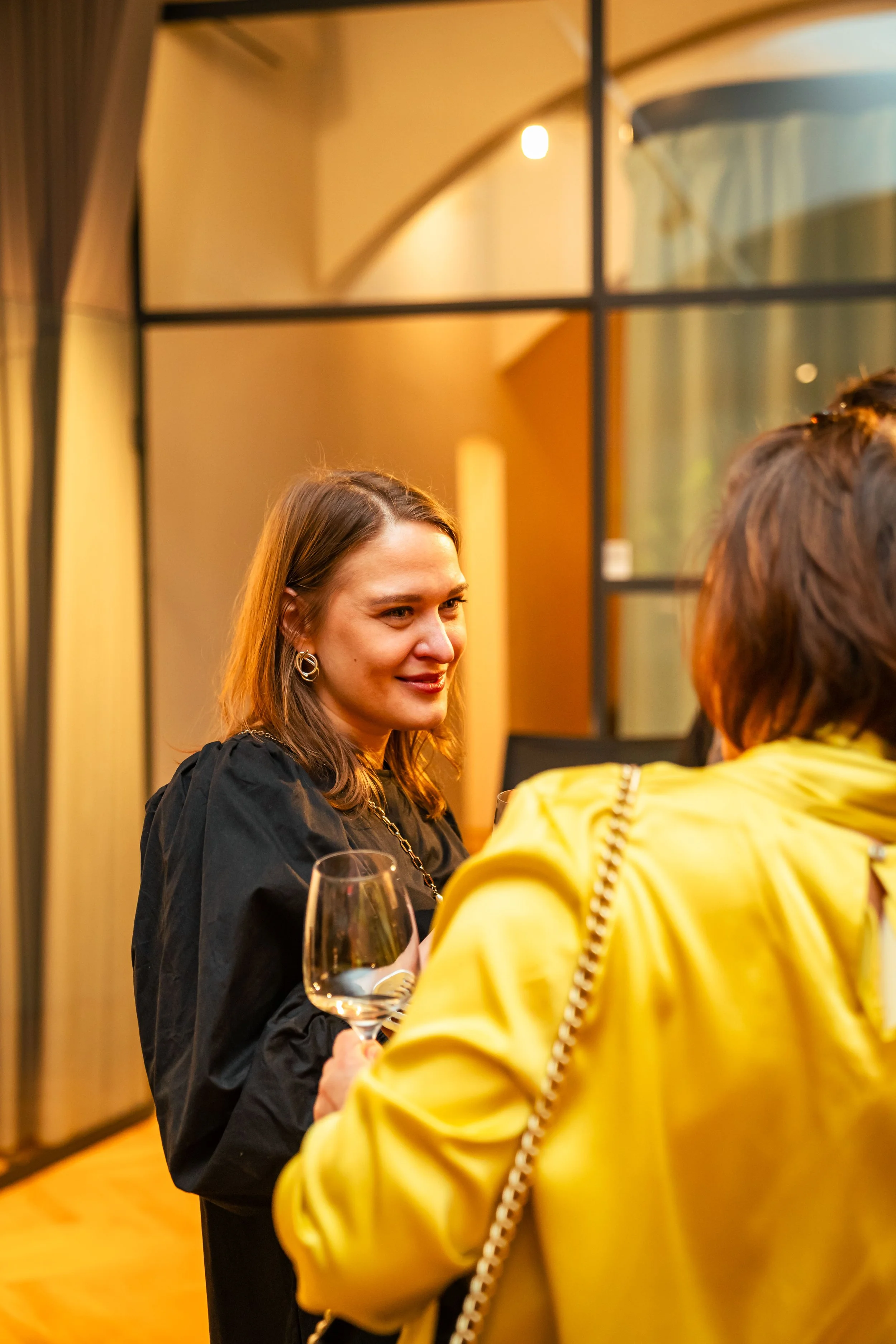 Zwei Frauen bei einem Gespräch in einem Restaurant, eine hält ein leeres Weinglas, die andere trägt eine gelbe Jacke, im Hintergrund große Fenster mit schwarzem Rahmen.