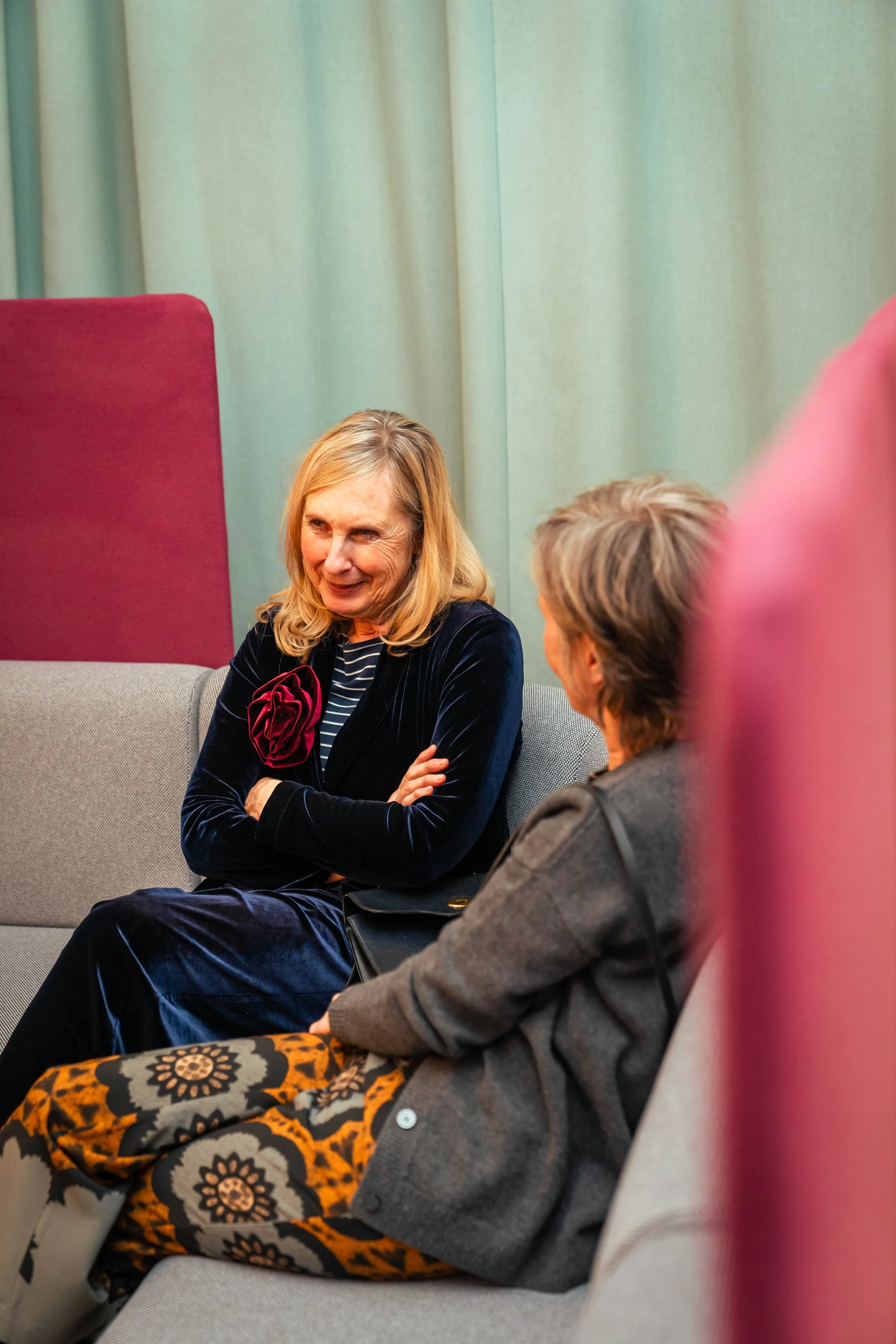 Zwei Frauen sitzen auf einer Couch und unterhalten sich. Eine Frau trägt ein dunkles Samtkleid mit rotem Blütenaccessoire, die andere trägt einen grauen Mantel.