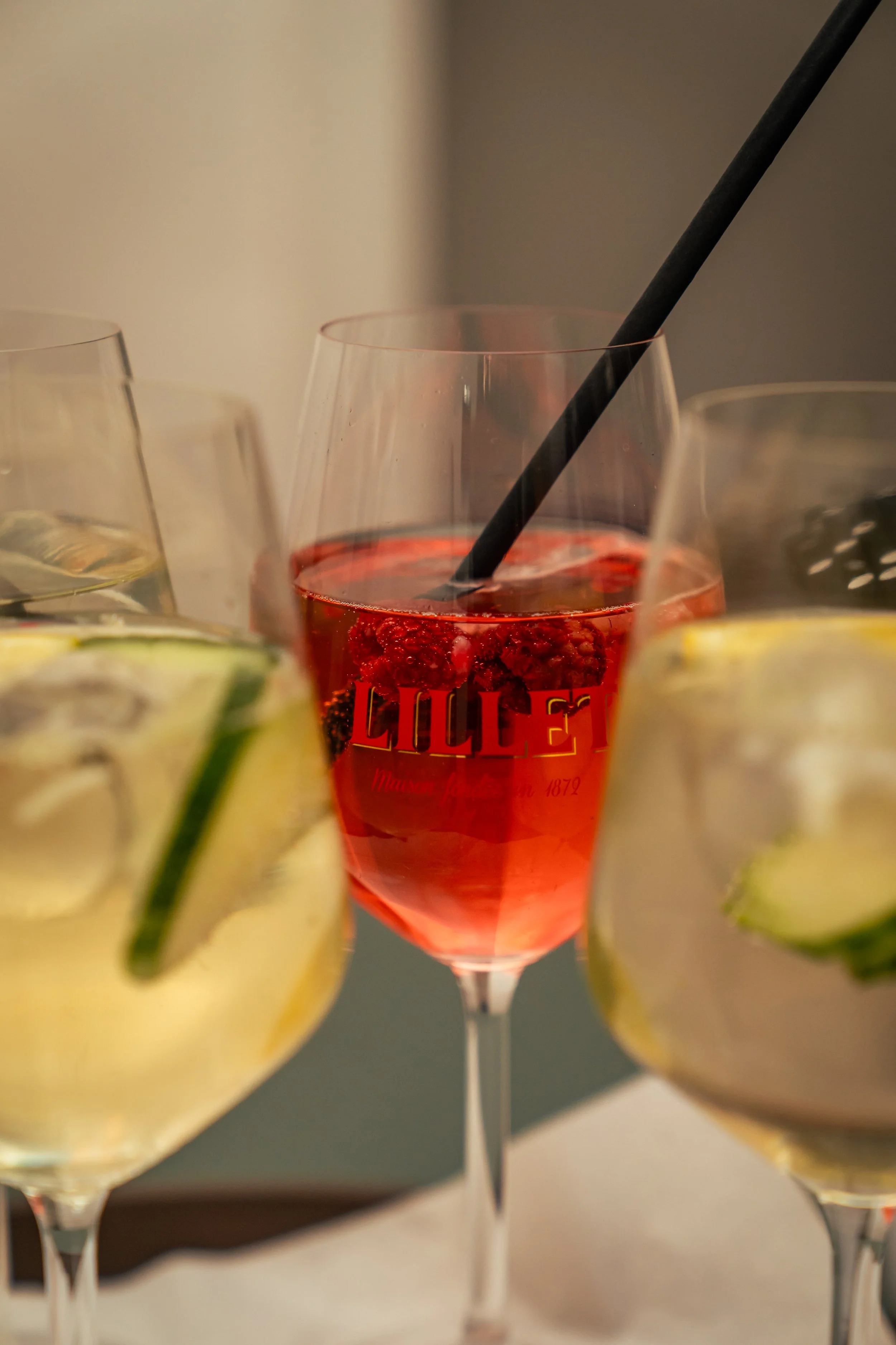 Mehrere Weingläser mit verschiedenen Aperitifs, darunter ein rotes Getränk in der Mitte, umrahmt von klaren Gläsern mit Zitronen- und Gurkenscheiben.