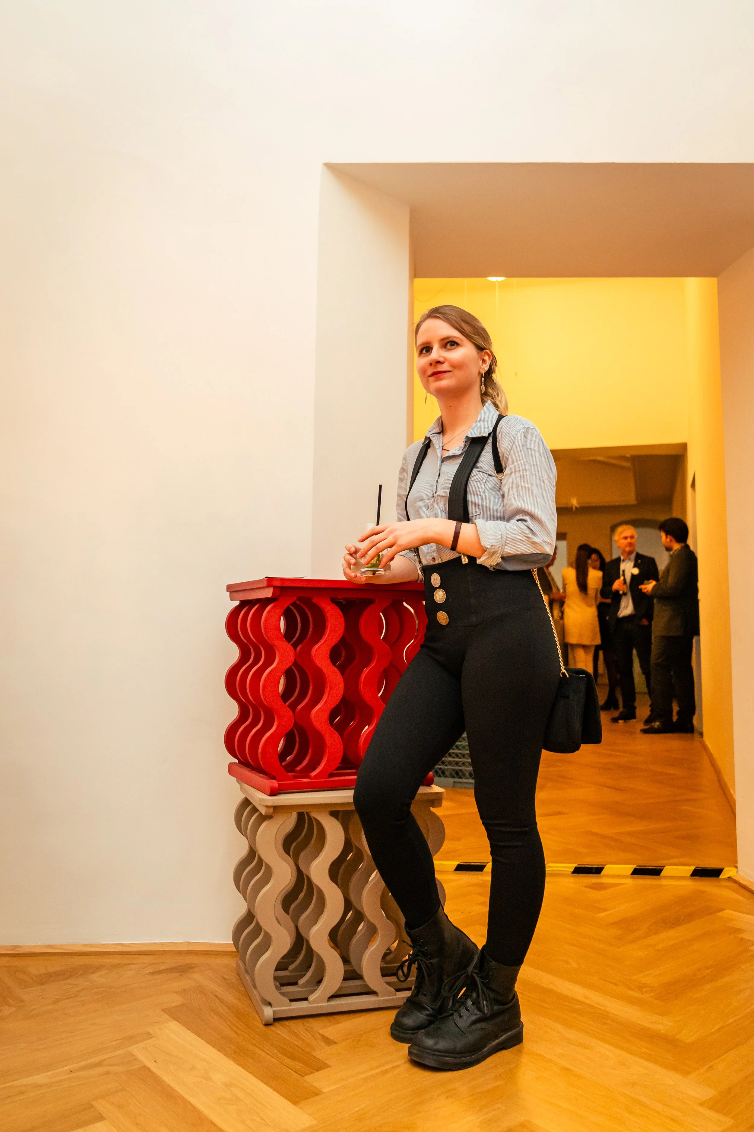 Junge Frau mit grauem Hemd, schwarzen Hosen und schwarzen Stiefeln steht in einer Galerie und hält ein Glas. Im Hintergrund sind weitere Personen bei einer Veranstaltung.