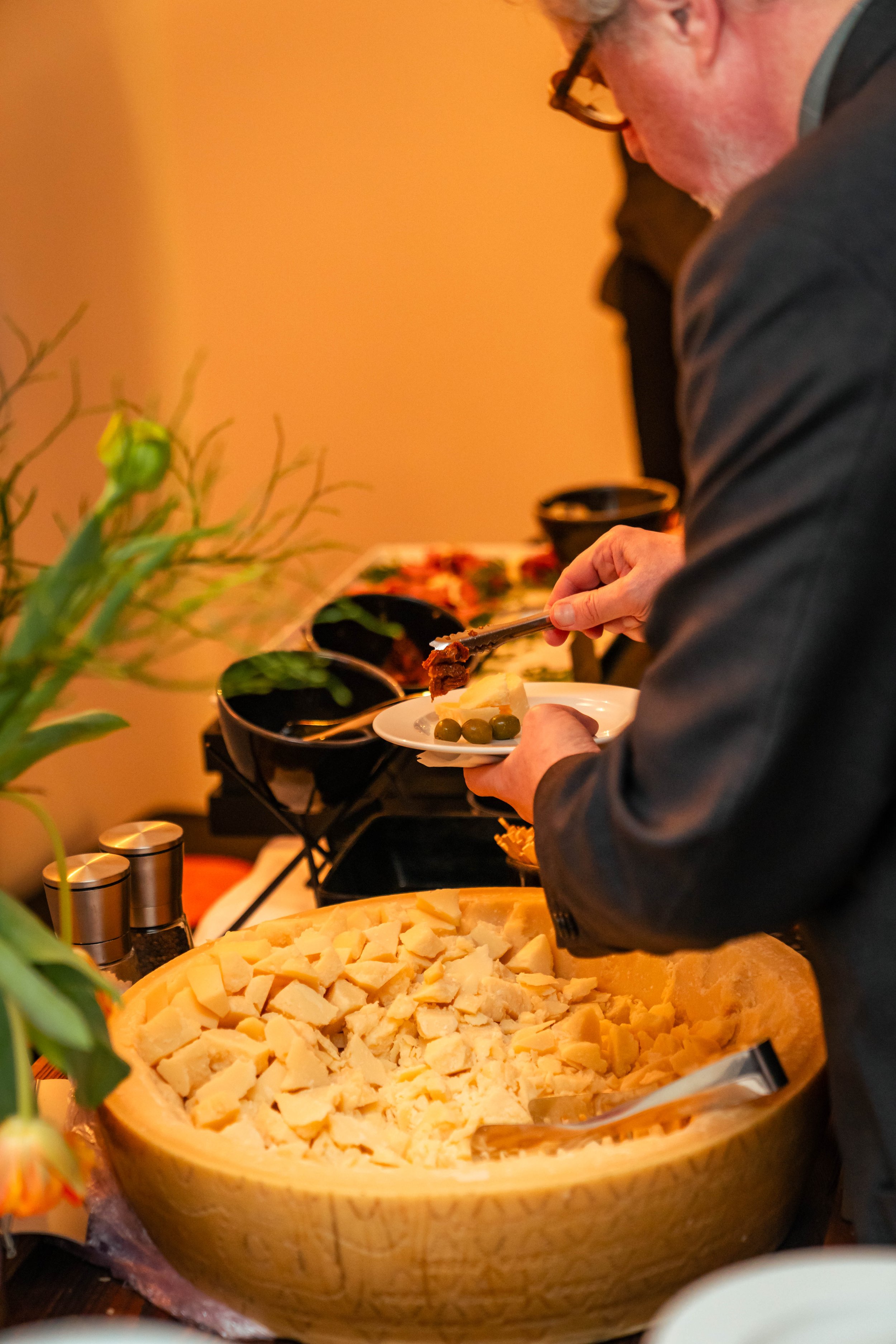 Person bedient sich bei einem Buffet mit verschiedenen Käsesorten, Gurken und weiteren Speisen.