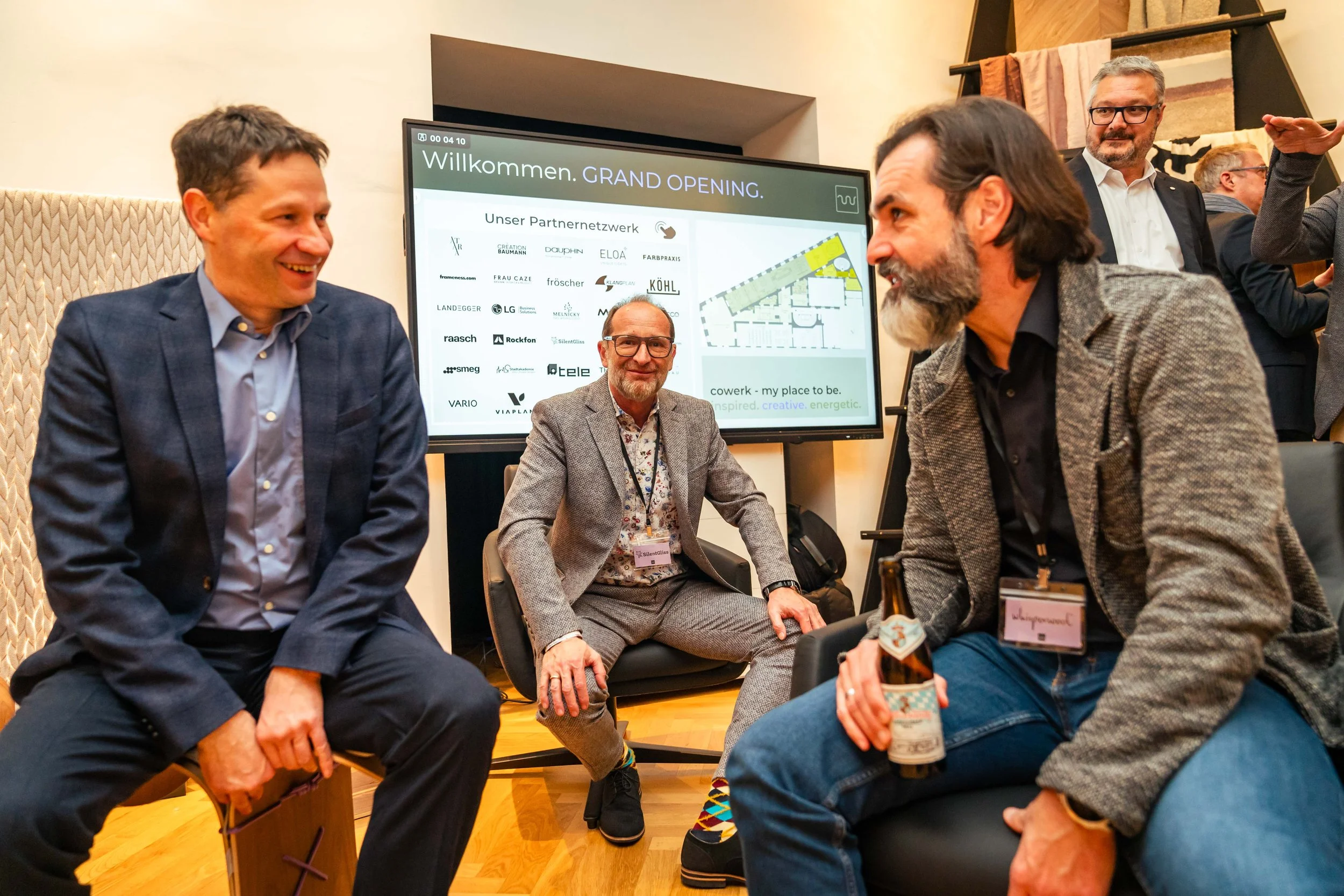 Drei Männer sitzen und lachen bei einem Networking-Event, im Hintergrund ein Bildschirm mit Willkommensgruß und Partnerlogos.