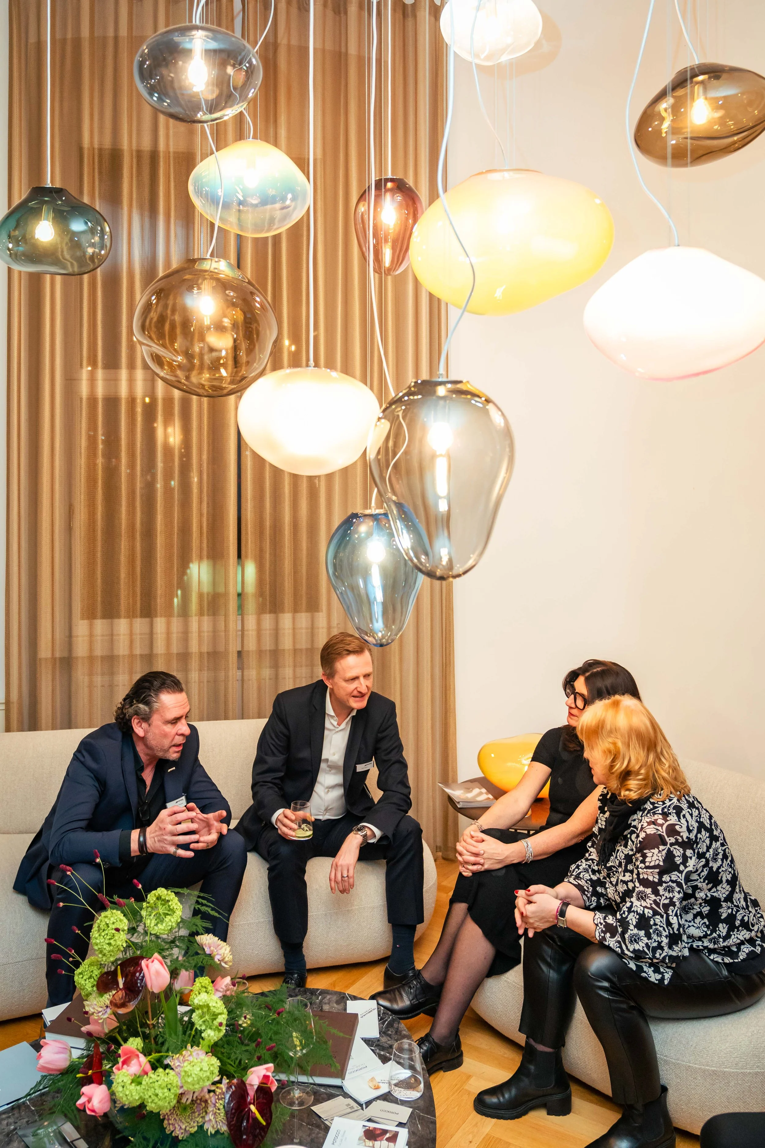 Vier Menschen sitzen auf einem Sofa, unter bunten modernen Lampen, im Gespräch bei einer Cocktailparty, mit Blumenstrauß auf einem Tisch davor.