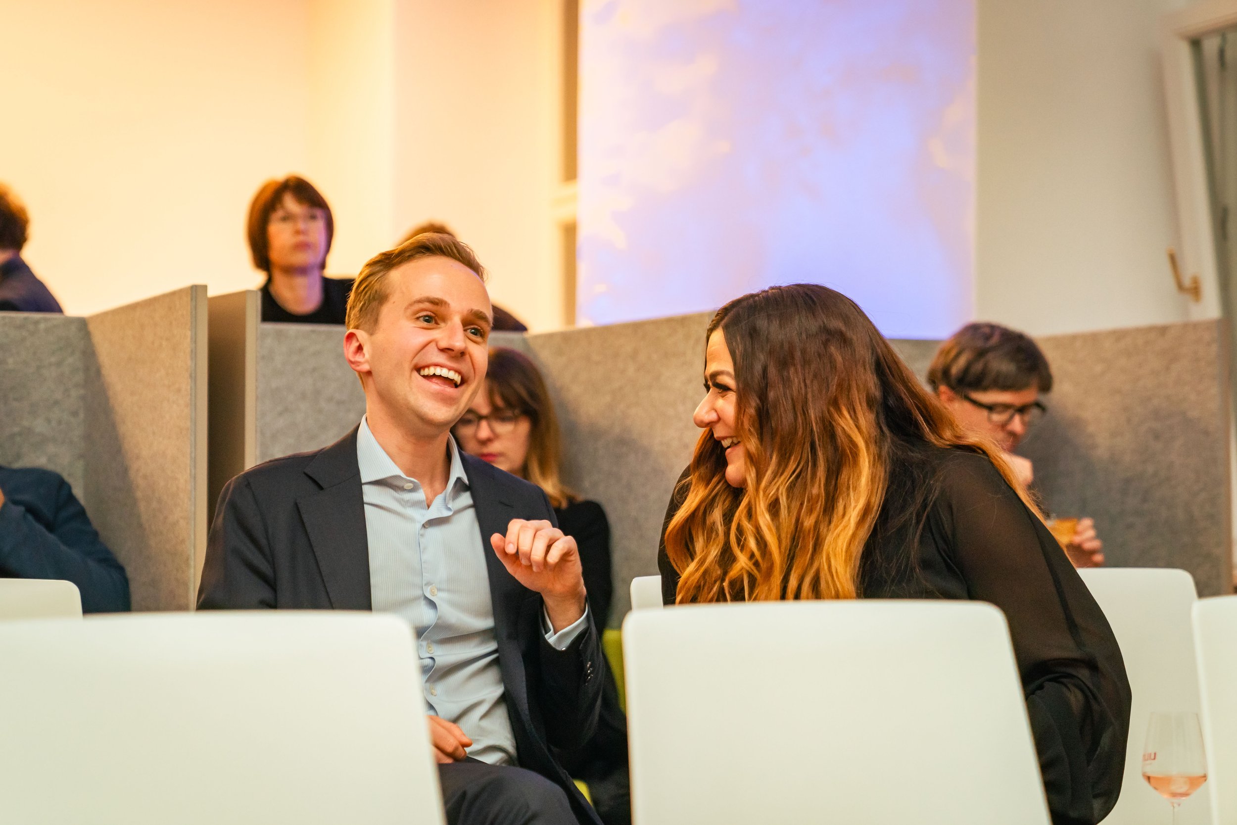 Zwei Menschen lachen und unterhalten sich bei einer Veranstaltung, im Hintergrund weitere Personen.