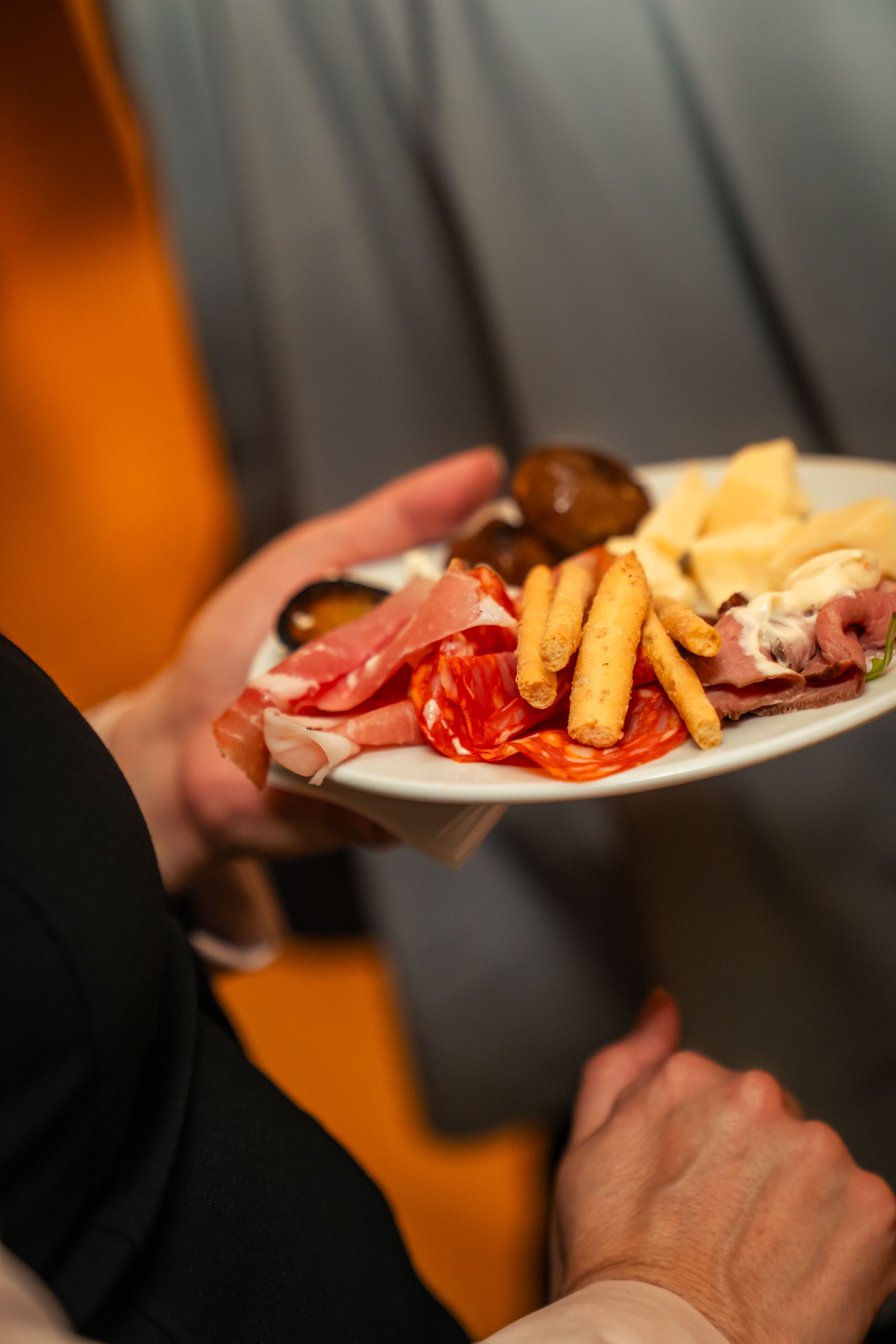 Eine Hand hält einen Teller mit verschiedenen mediterranen Antipasti, darunter Schinken, Salami, Grüne Oliven, Käse, Taco Chips und Fleischsalat.