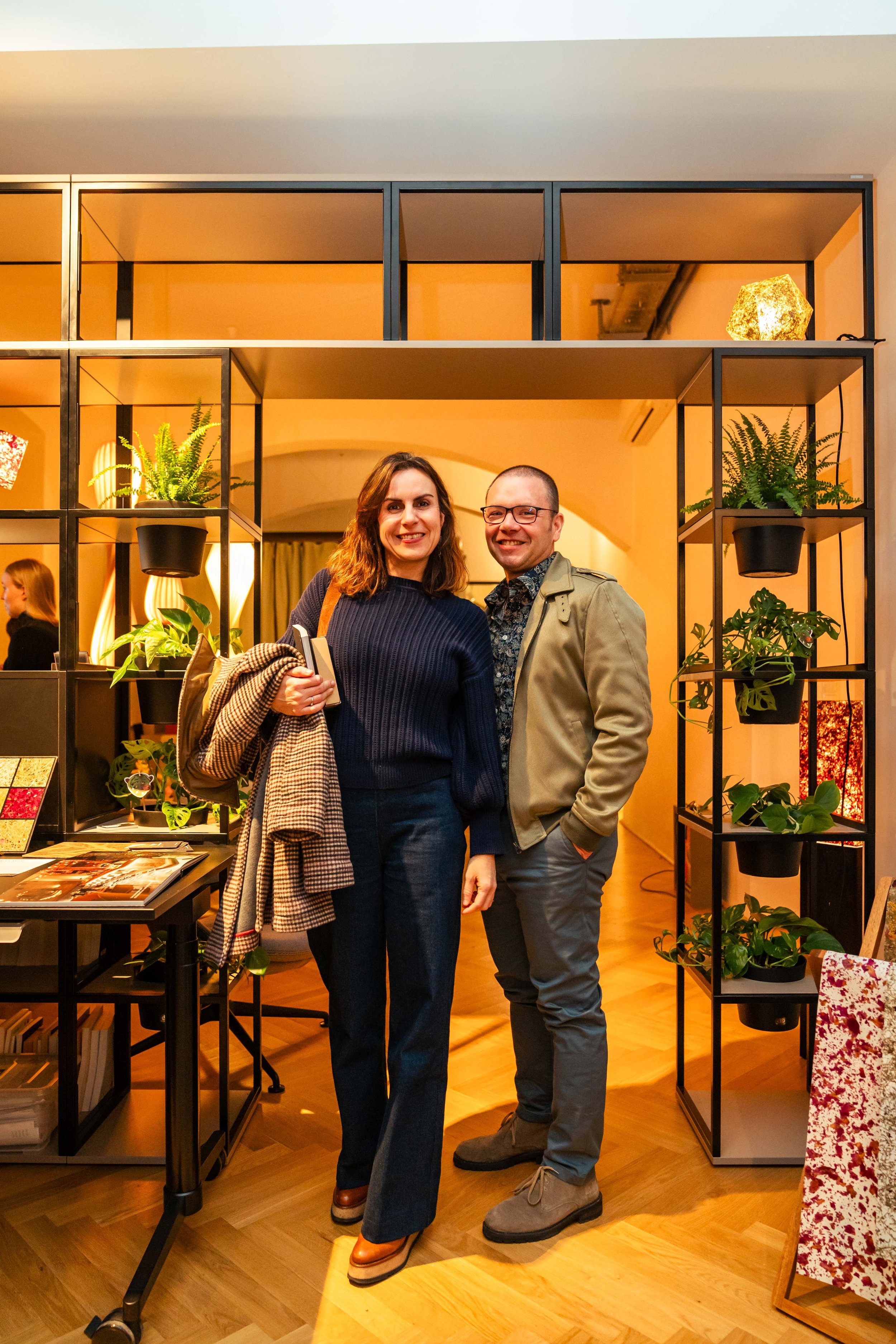 Zwei Menschen, eine Frau und ein Mann, stehen vor einer Regalkonstruktion mit Pflanzen in einem Raum mit warmem Licht. Die Frau trägt eine dunkelblaue Pullover und dunkle Jeans, die Frau trägt eine beige Jacke, ein Hemd mit Blumenmuster und graue Hos