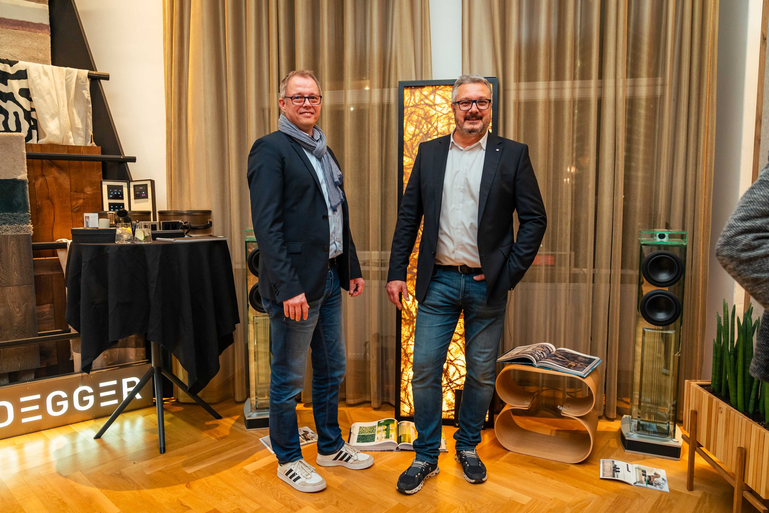 Zwei Männer in Anzügen stehen in einem Wohnzimmer mit Holzfußboden, hinter ihnen einen Bildschirm mit einem Herbstbild, links eine Stehlampe und Tonanlage, rechts eine Pflanze und ein offener Zeitschriftenständer.