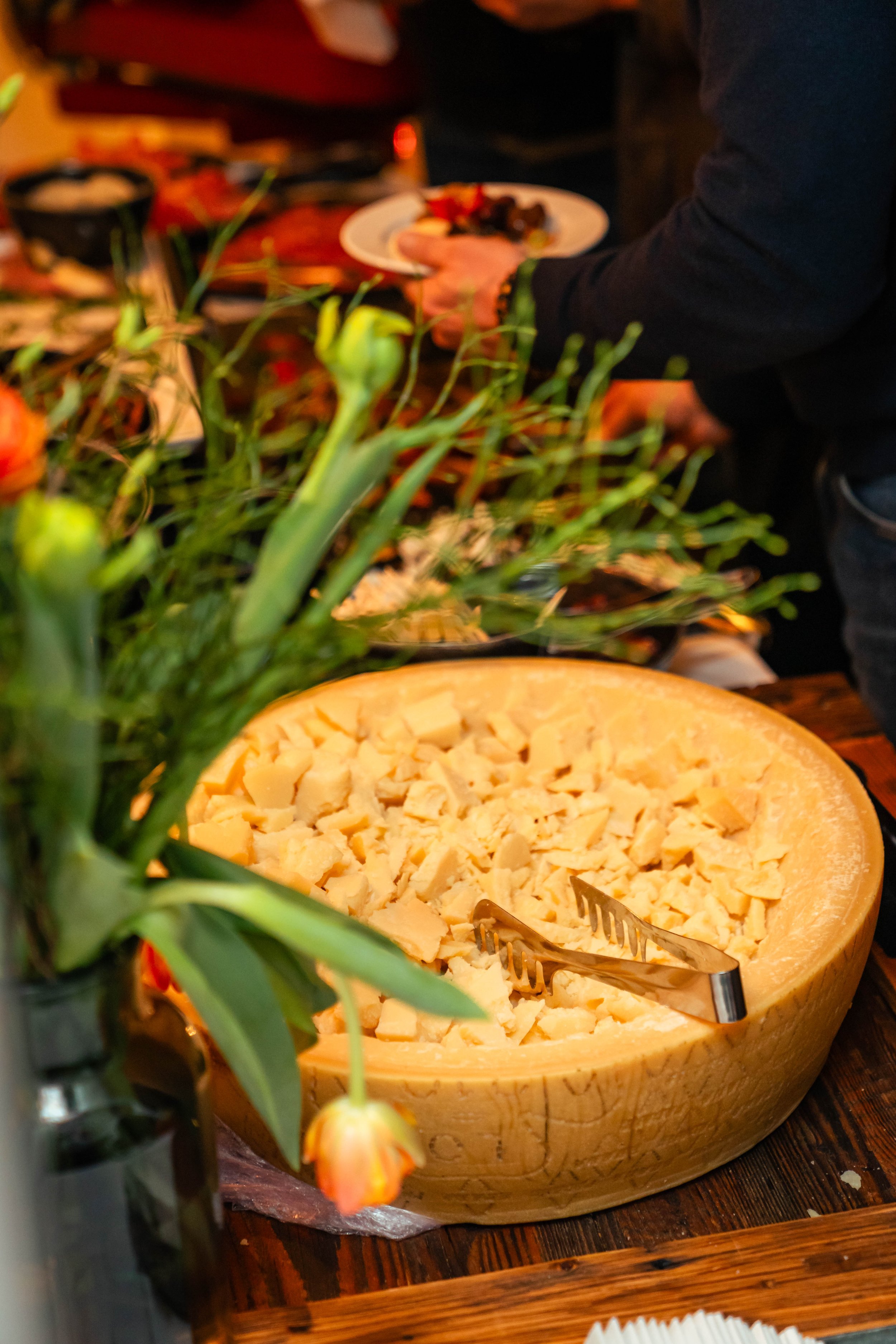 Großer Käseblock mit Käsemessern auf einem Holztisch, im Vordergrund Tafel mit Blumen