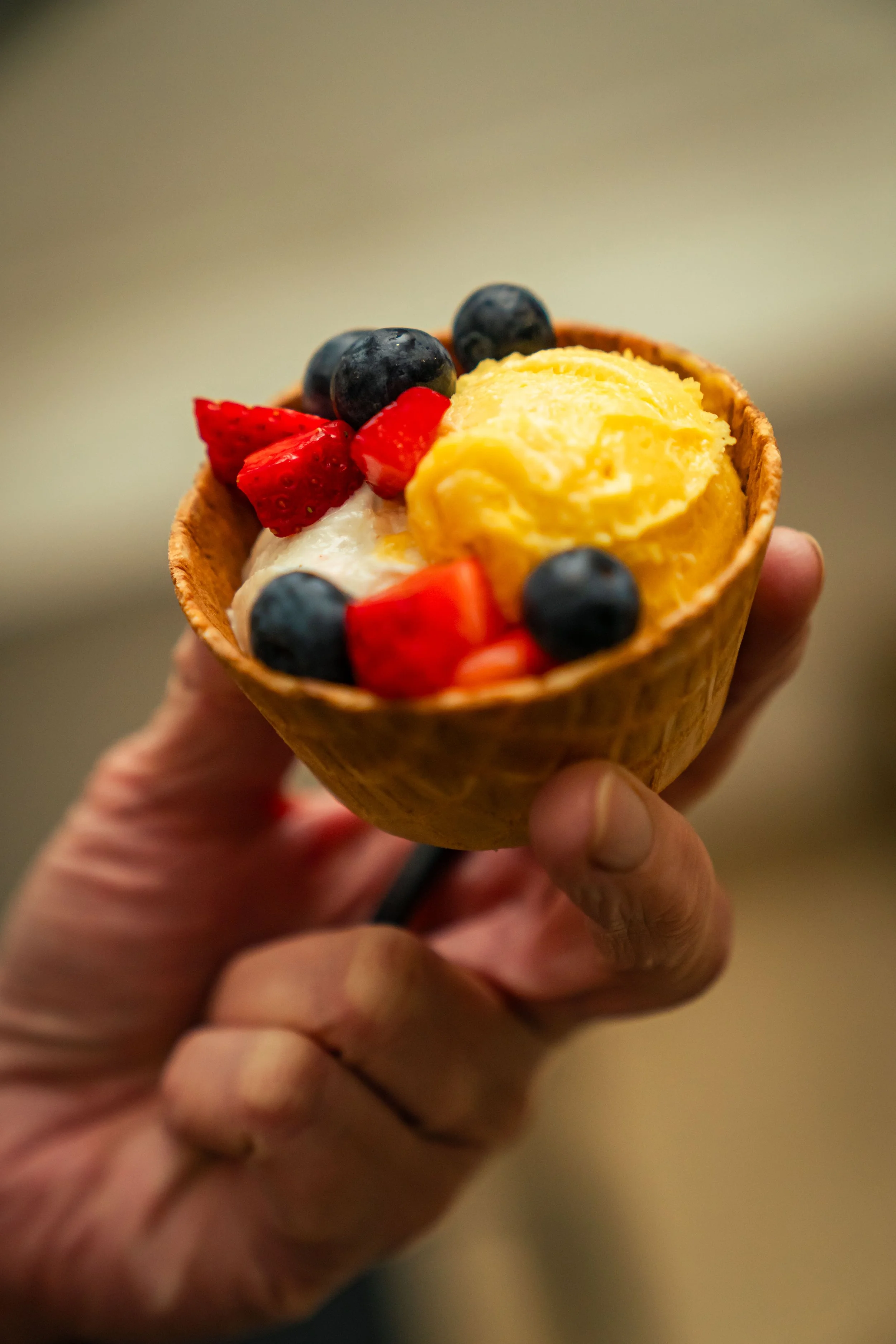 Hand hält eine Waffel mit Vanilleeis, frischen Erdbeeren, Heidelbeeren und einer Kugel Mango-Eis.