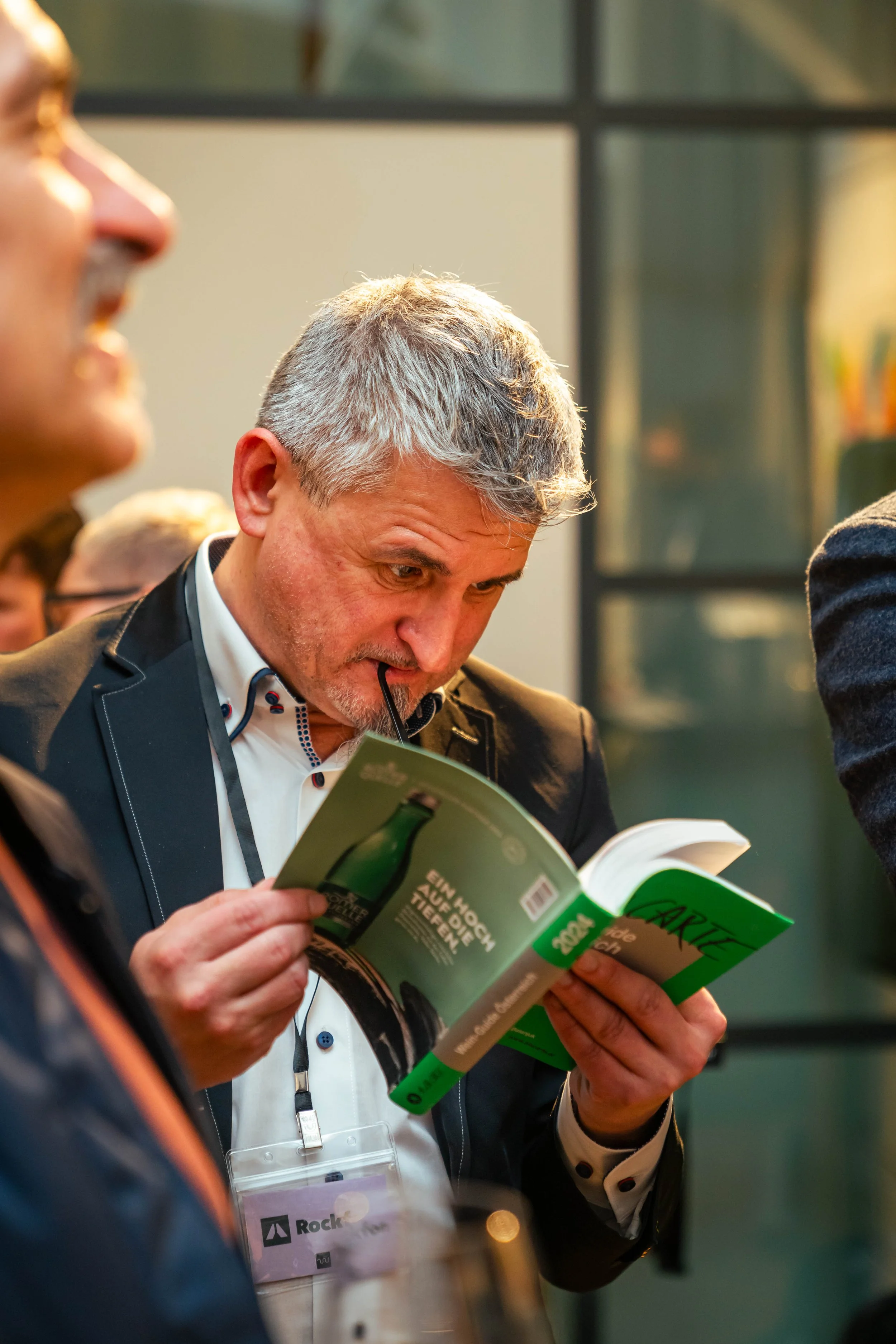 Ein Mann liest ein Buch auf einer Veranstaltung, während er eine Brille zwischen den Lippen hält. Neben ihm sind andere Personen zu sehen, und er trägt eine Namensschleife.