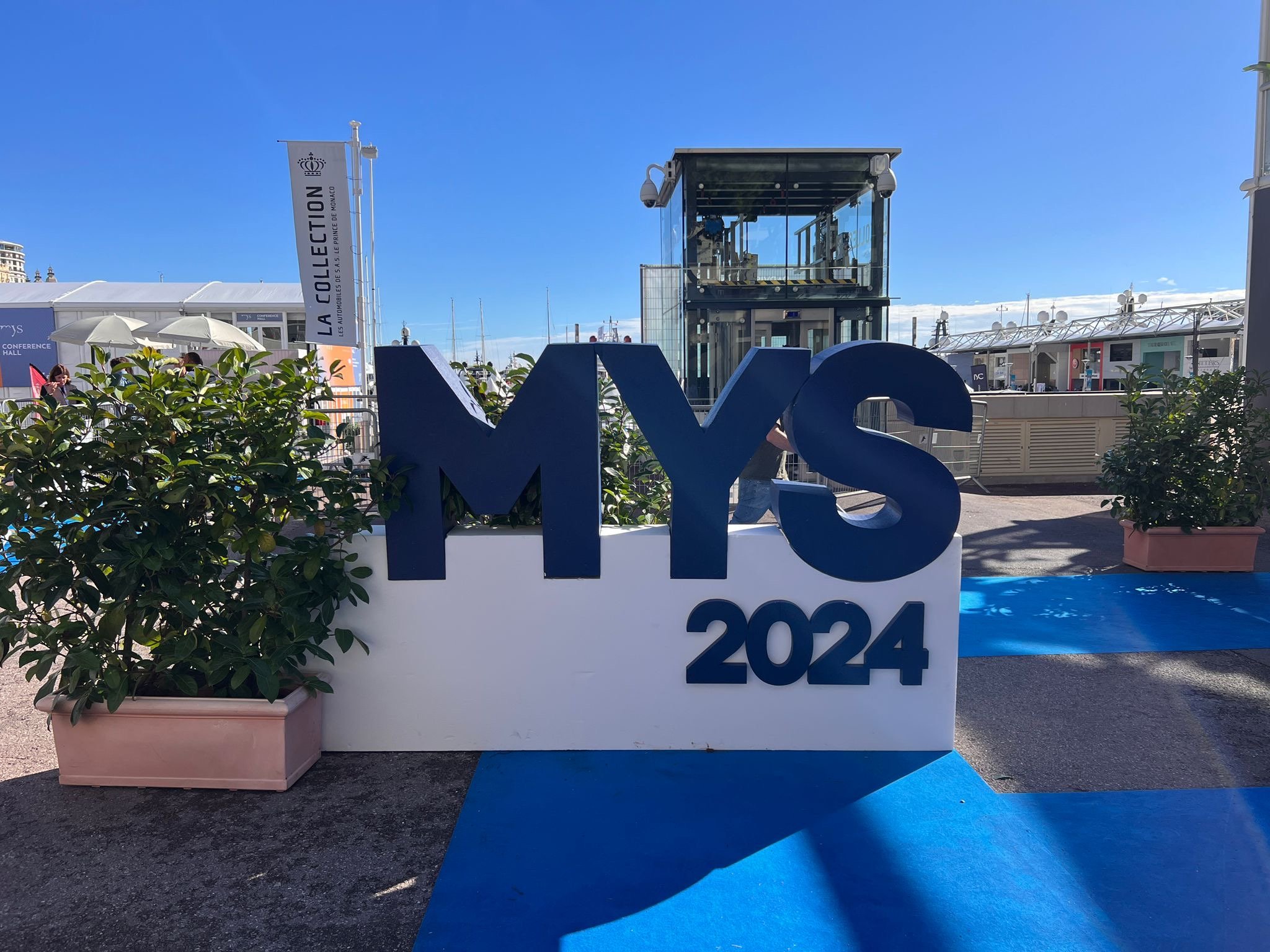 Große blaue Buchstaben und Zahlen mit der Aufschrift 'MYS 2024' auf einem weißen Hintergrund, im Hintergrund ein Dock mit Yachten, Sonnenschirme und Menschen, blauer Himmel.