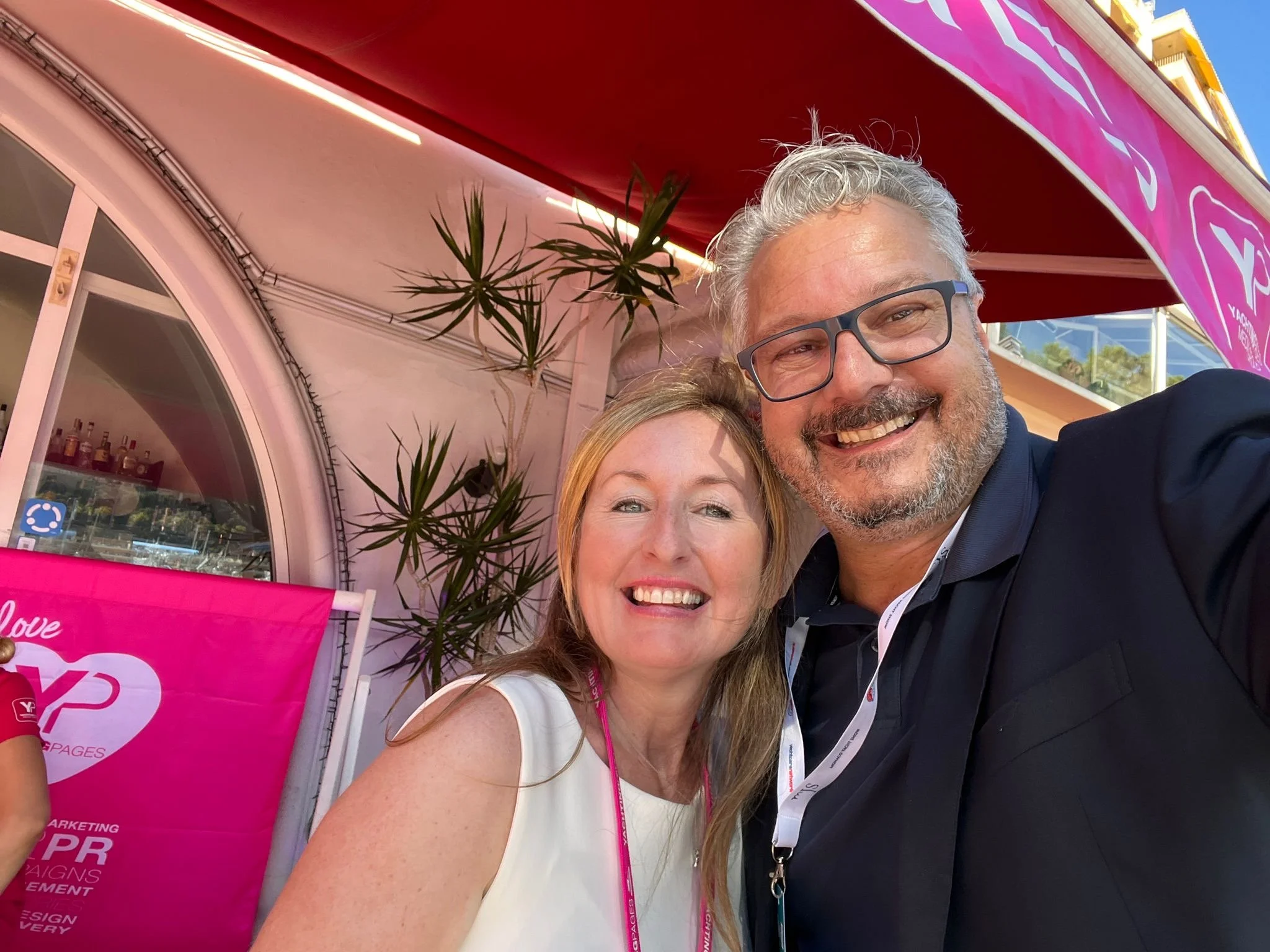 Ein Mann und eine Frau posieren lachend für ein Selfie vor einem pinken Stand mit Pflanzen im Hintergrund.