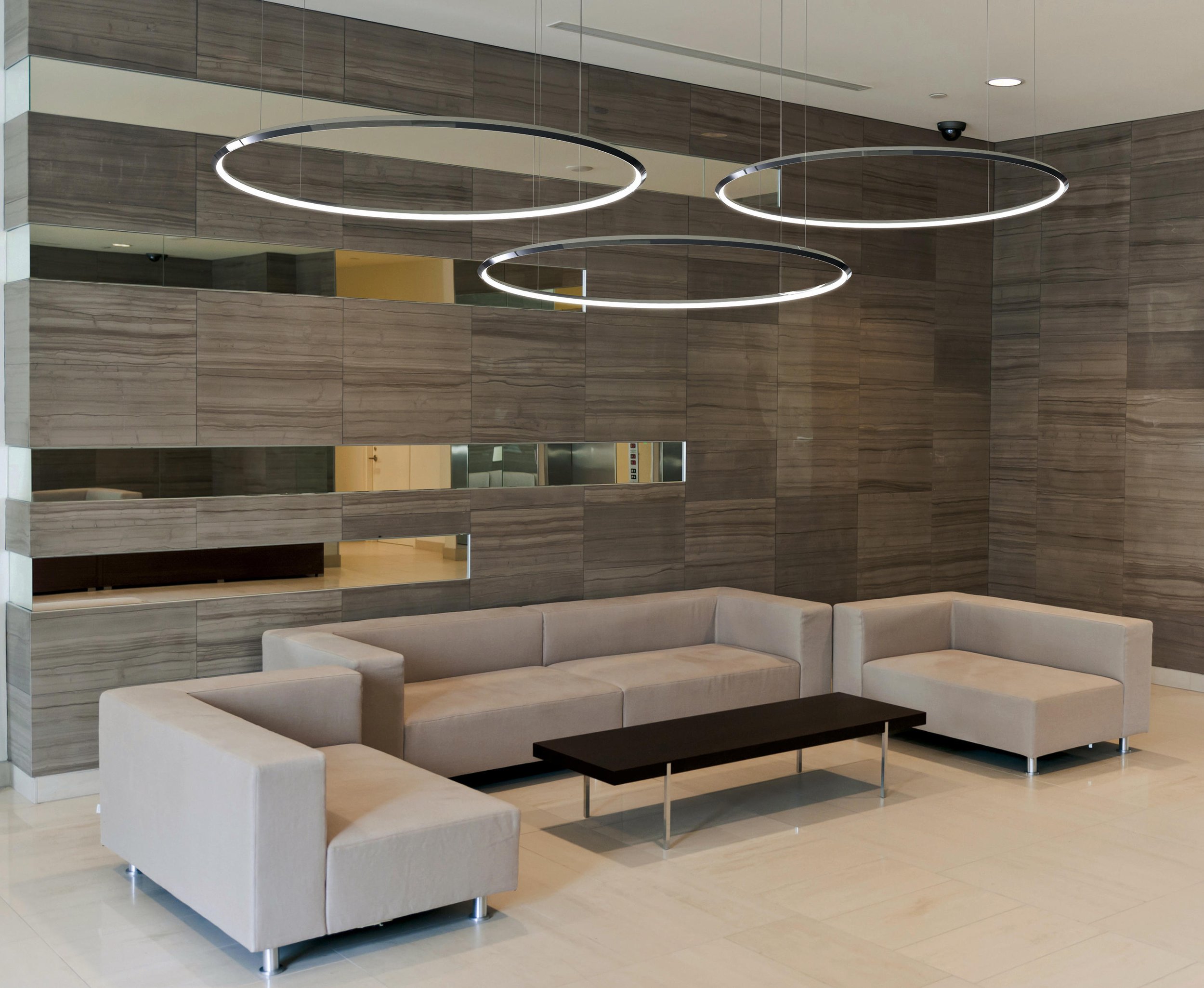 Moderne Lobby mit grauem Sofa, braunem Couchtisch, dunklen Holzpaneelen an der Wand und drei runden LED-Leuchten an der Decke.