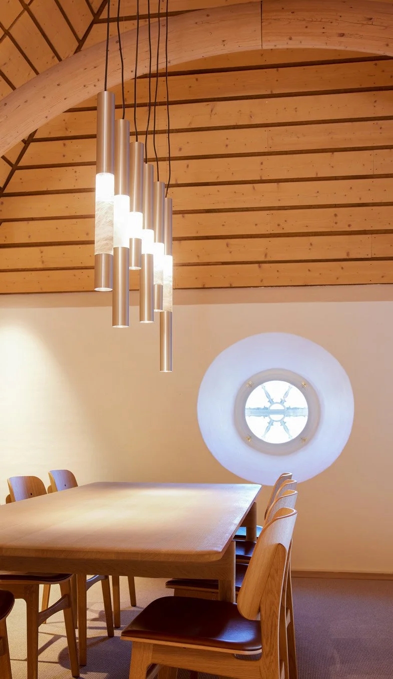 Innenraum eines Raumes mit Holzdecke, einem langen Holztisch, Holzhockern und modernen Hängeleuchten. Es gibt auch ein rundes Fenster in der weißen Wand.