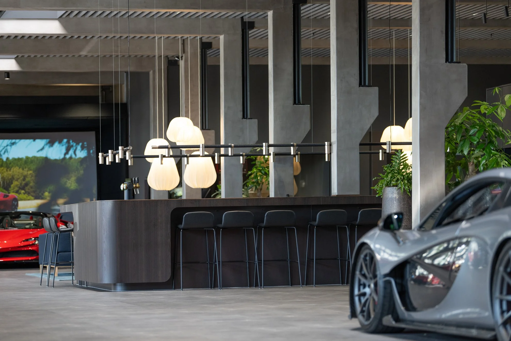 Innenraum eines modernen Autohauses mit hochwertigen Sportwagen, einer Bar mit Hockern, modernem Lichtdesign und Grünpflanzen.