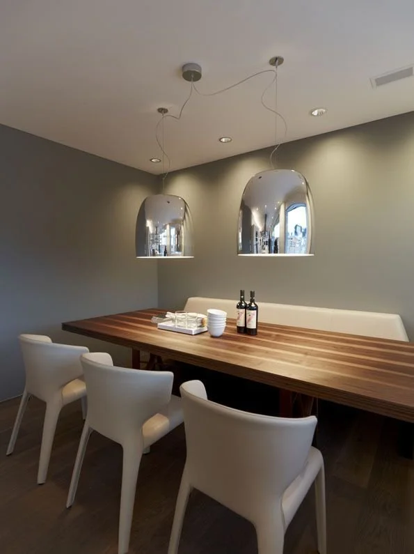 Moderner Essbereich mit einem langen Holztisch, weißen Stühlen und zwei silbernen Hängeleuchten. Die Wand ist hellgrau, und auf dem Tisch stehen Teller sowie Getränke.