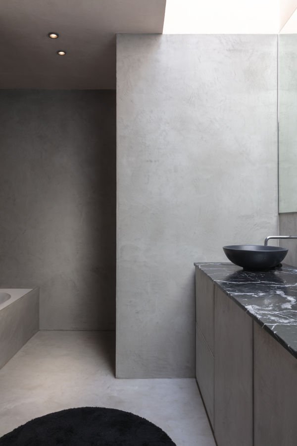 Modernes Badezimmer mit grauen Wänden, schwarzer Waschtrog auf schwarzer Marmorplatte, dunklem runden Teppich im Vordergrund, weiße Decke mit Einbauleuchten.