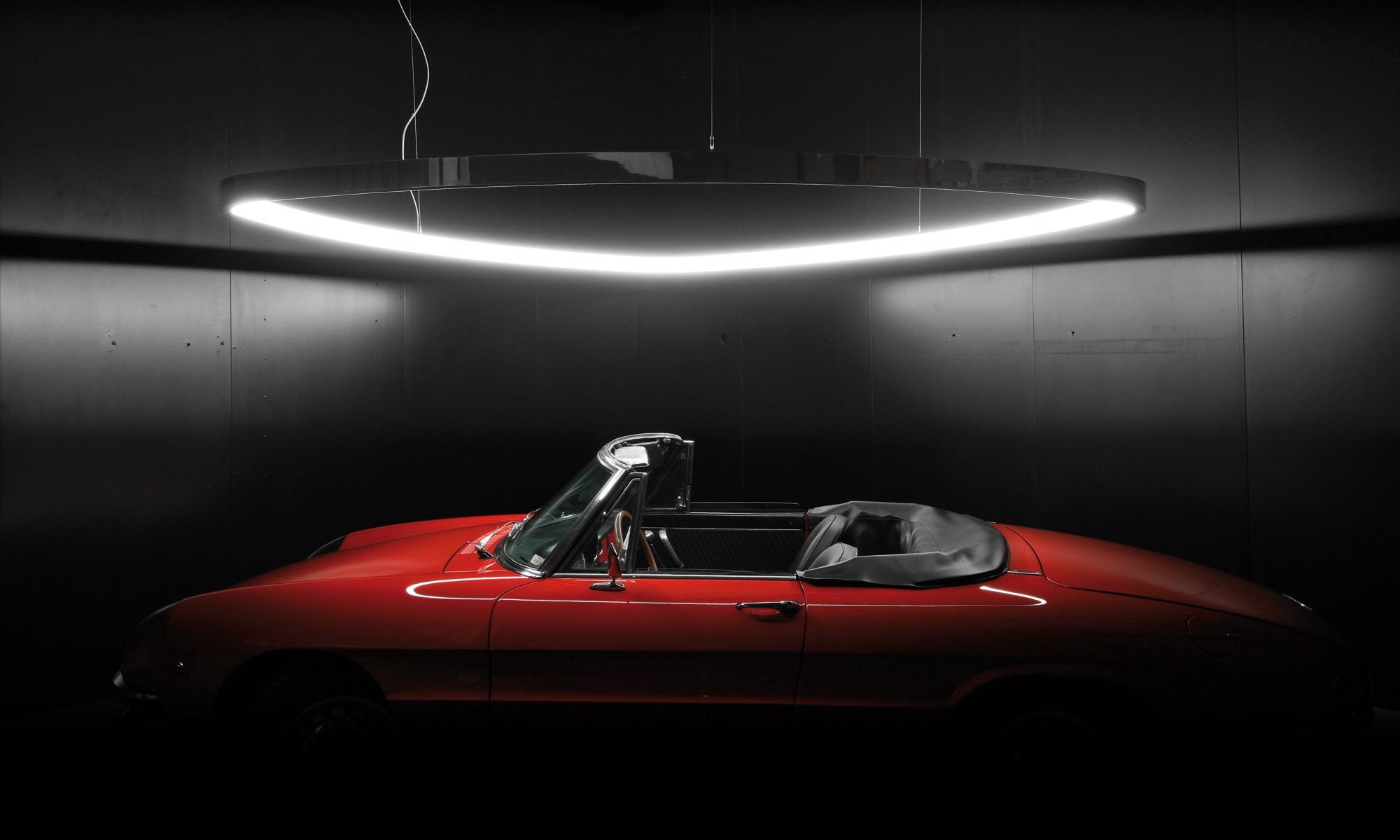 Roter Sportwagen in einem dunklen Raum unter einer ovalen Lichtinstallation.