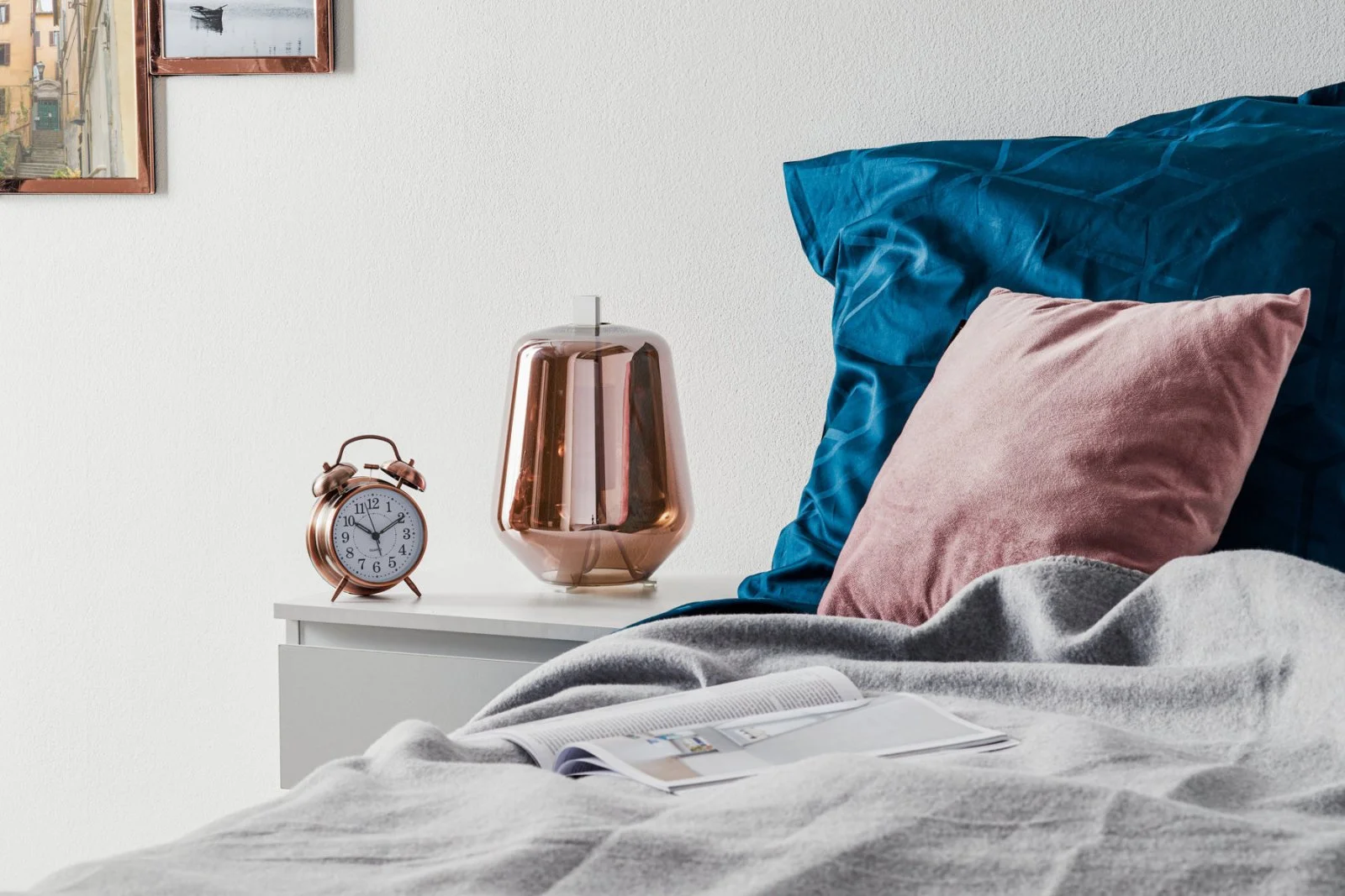 Schlafzimmer mit Bett, grauem Bettzeug, blauen und rosa Kissen, einer geöffneten Zeitschrift, Nachttisch mit Rosegoldfarbenem Wecker und passender Nachttischlampe, Wandbilder