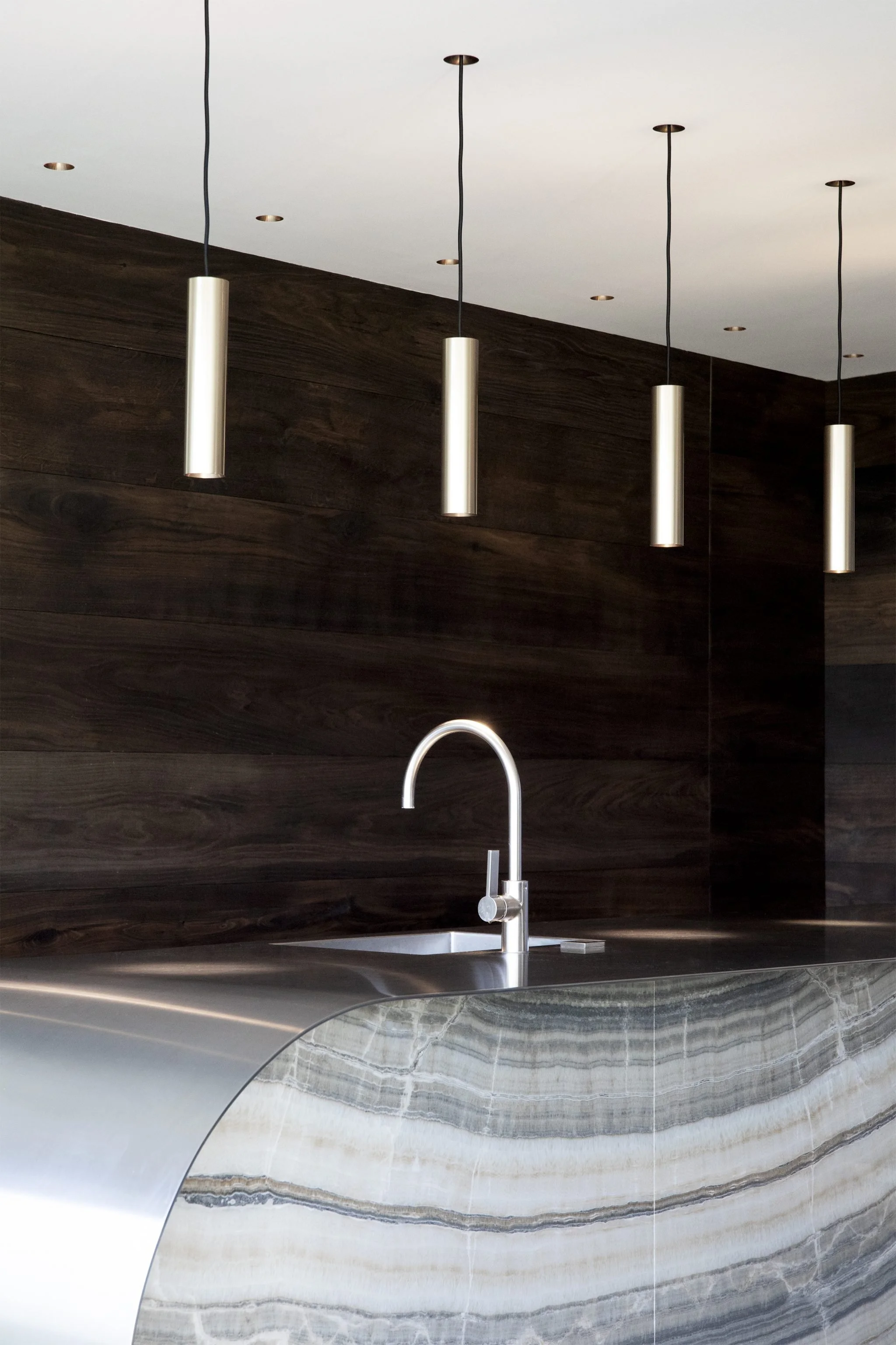 Moderne Küche mit dunkler Holzvertäfelung, einem Edelstahl-Spültisch mit Wasserhahn, und vier hängenden, zylindrischen Leuchten.
