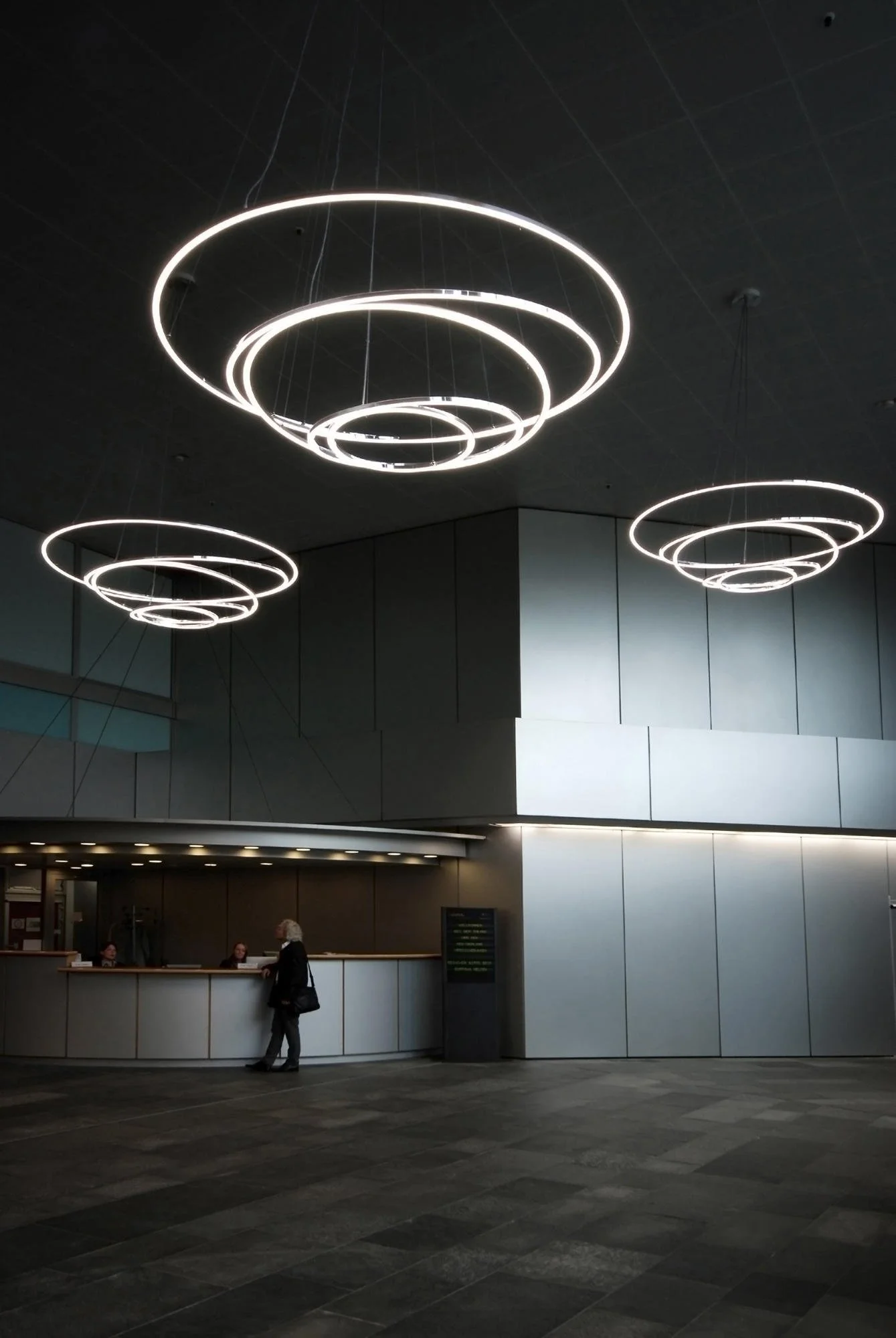 Innenaufnahme eines modernen Gebäudes mit dunklem Decke und drei großen, leuchtenden, kreisförmigen Lüftungskuppeln oder Lichtinstallationen. Im Vordergrund befindet sich ein Empfangstresen, an dem eine ältere Frau mit schwarzen Oberteil und schwarze