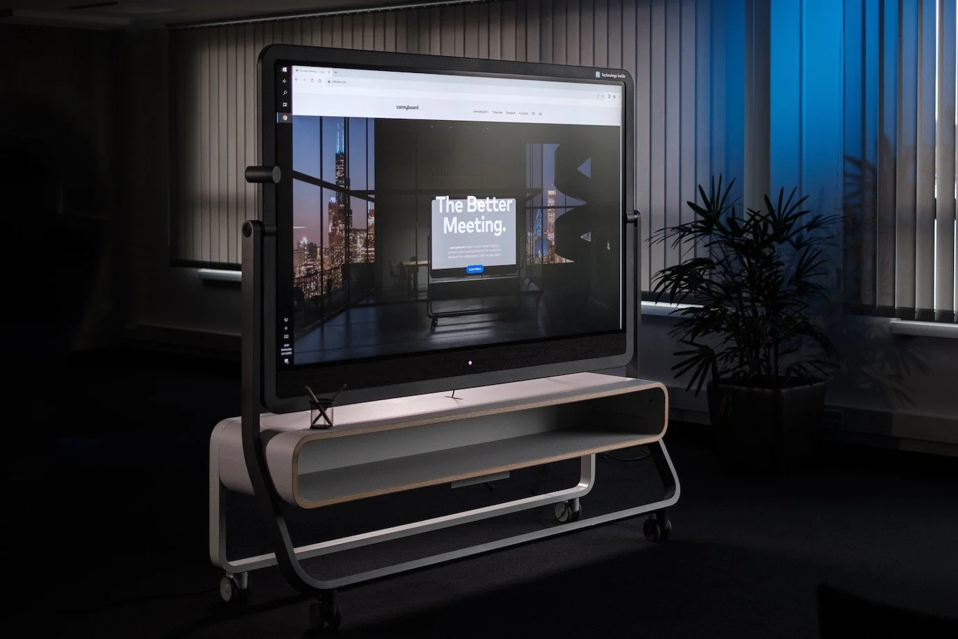 Großer Bildschirm mit Stadtansicht im Hintergrund, displaying den Text "The Better Meeting", in einem modernen, dunklen Raum mit Pflanzen und vertikalen Fensterjalousien.