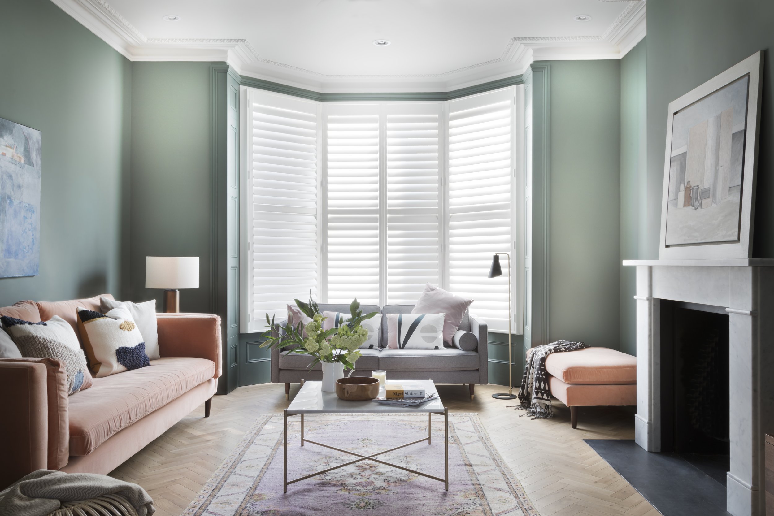 Gemütliches Wohnzimmer mit grünen Wänden, großen Fenstern mit weißen Jalousien, gepolstertem Sofa in Peach, einem kleineren grauen Sofa, einem weißen Couchtisch mit Blumen, einem Kamin und moderner Kunst an den Wänden.