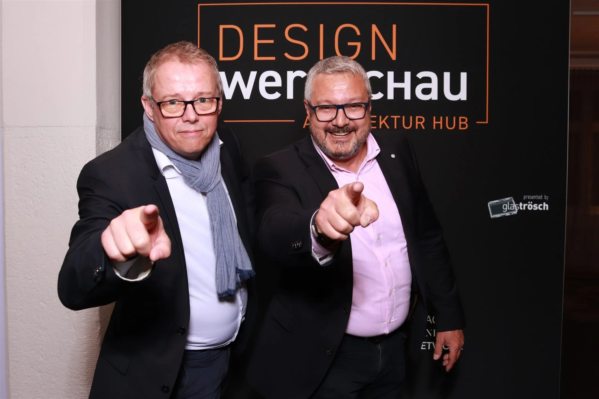 Zwei Männer in formeller Kleidung zeigen mit Fingern auf die Kamera vor einem schwarzen Hintergrund mit dem Text 'Design Power Schau'.