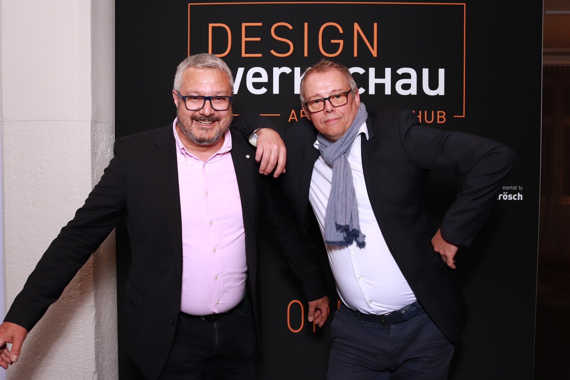 Zwei Männer in formeller Kleidung posieren lachend vor einem schwarzen Hintergrund mit orange und weißen Text, der ‚DESIGN‘ und ‚VERNISSAGE‘ erwähnt. Einer hat graues Haar und trägt eine Brille, der andere trägt eine Brille, eine schwarze Jacke, ein weißes Hemd und einen grauen Schal.