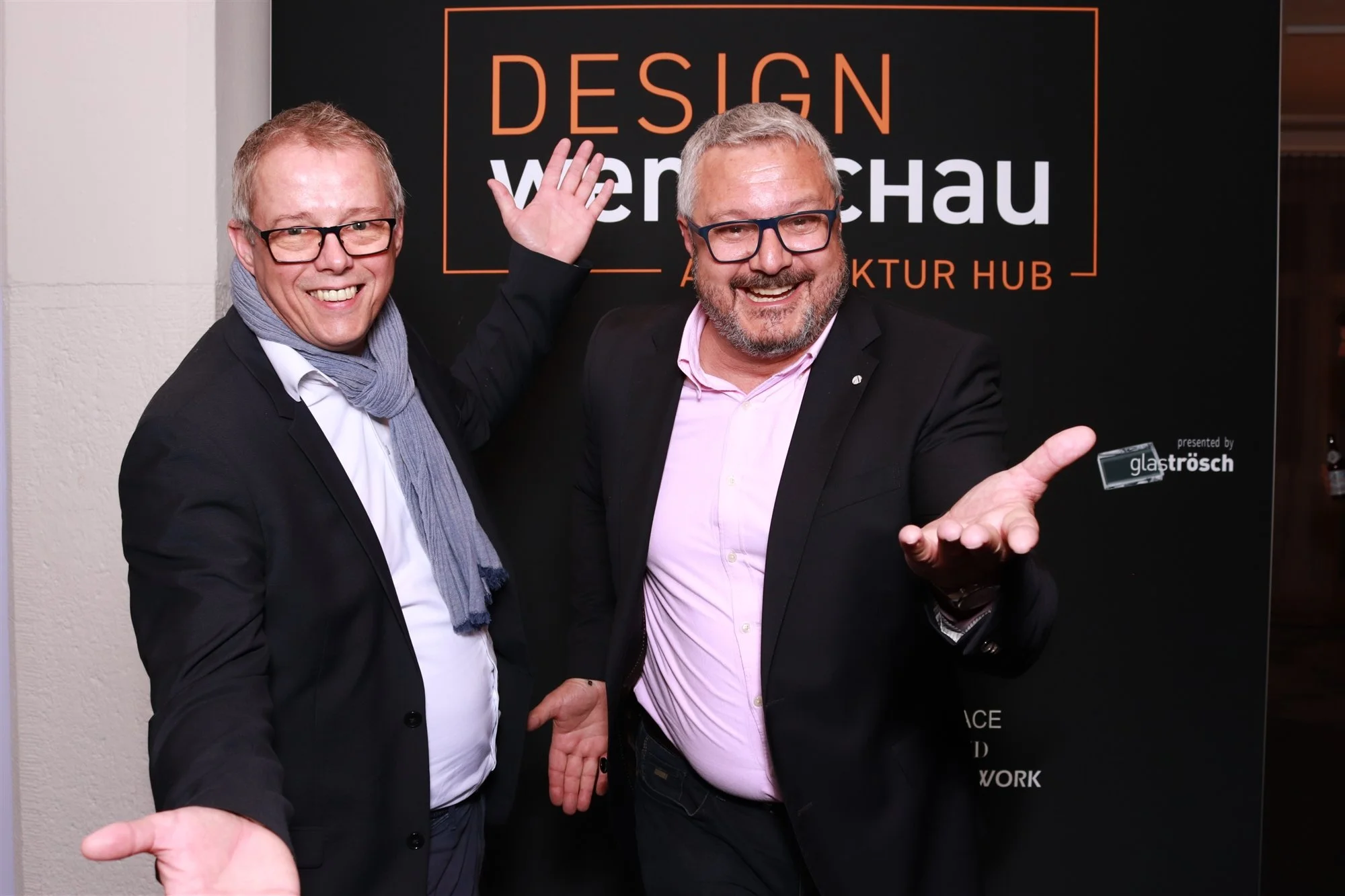 Zwei lachende Männer in Anzügen vor einem schwarzen Banner mit orange und weißen Texten, die 'Design Wire Schau' und 'Architektur Hub' sagen. Einer der Männer hat eine Brille, trägt eine dunkle Jacke und einen rosa Hemd, der andere trägt ebenfalls eine Brille, eine dunkle Jacke und einen grauen Schal.