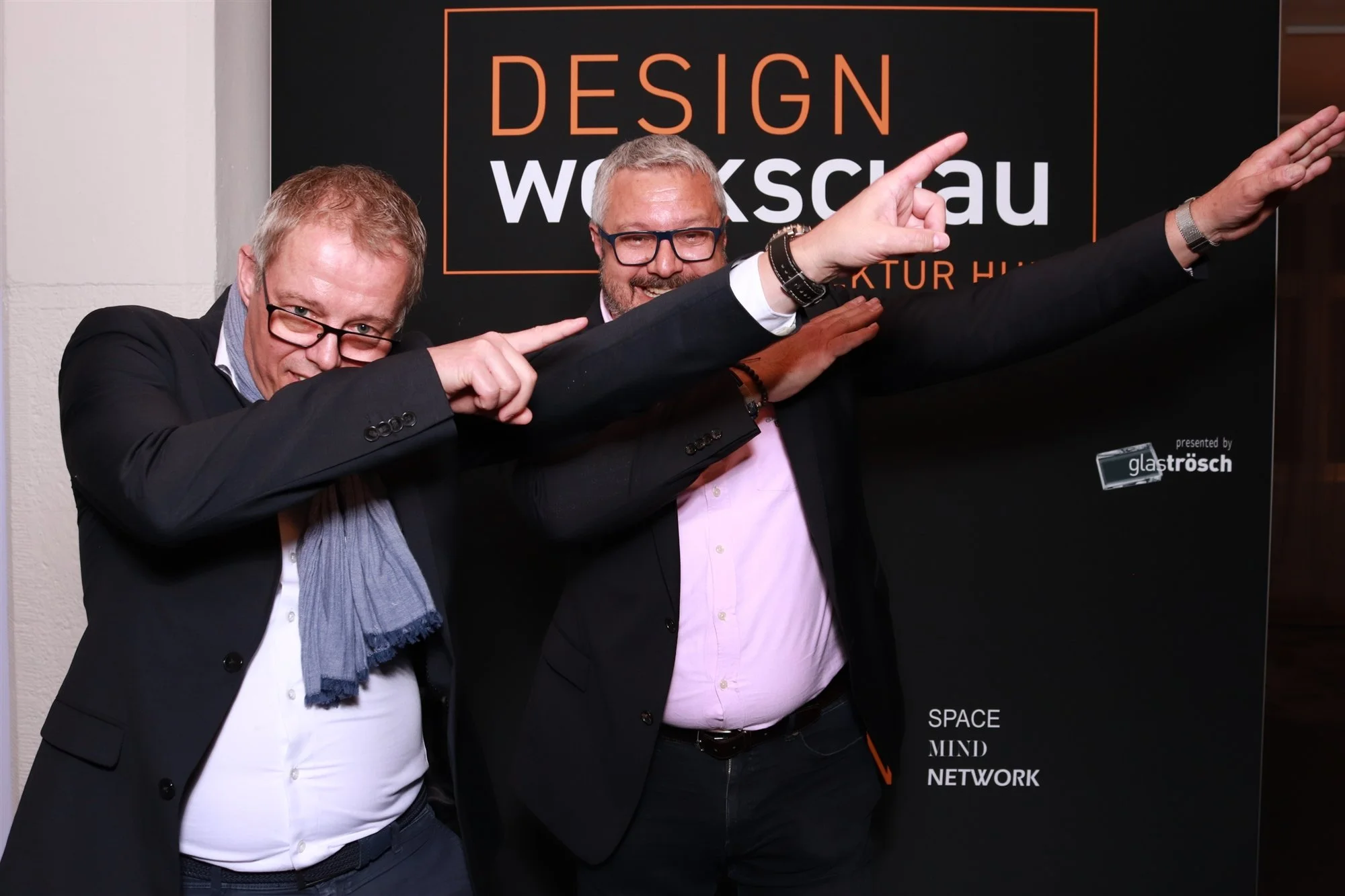 Zwei Männer in Businesskleidung und Brillen machen eine Pose vor einem schwarzen Banner mit orangefarbenem Text. Der Text auf dem Banner lautet 'DESIGN WOKSKAU' und weitere kleinere Wörter darunter, darunter 'SPACE', 'MIND', 'NETWORK'. Der Mann auf der linken Seite trägt einen grauen Schal. Beide lachen und zeigen eine Armbewegung, die nach oben zeigt.