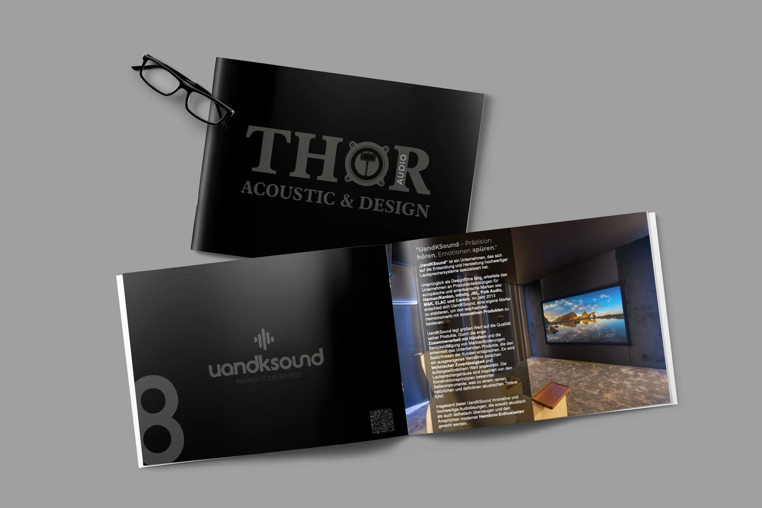 Gedrucktes Broschüren-Layout für Thor Acoustic & Design, geöffnet, mit Text, Bild und QR-Code, auf grauem Hintergrund, daneben schwarze Brille.