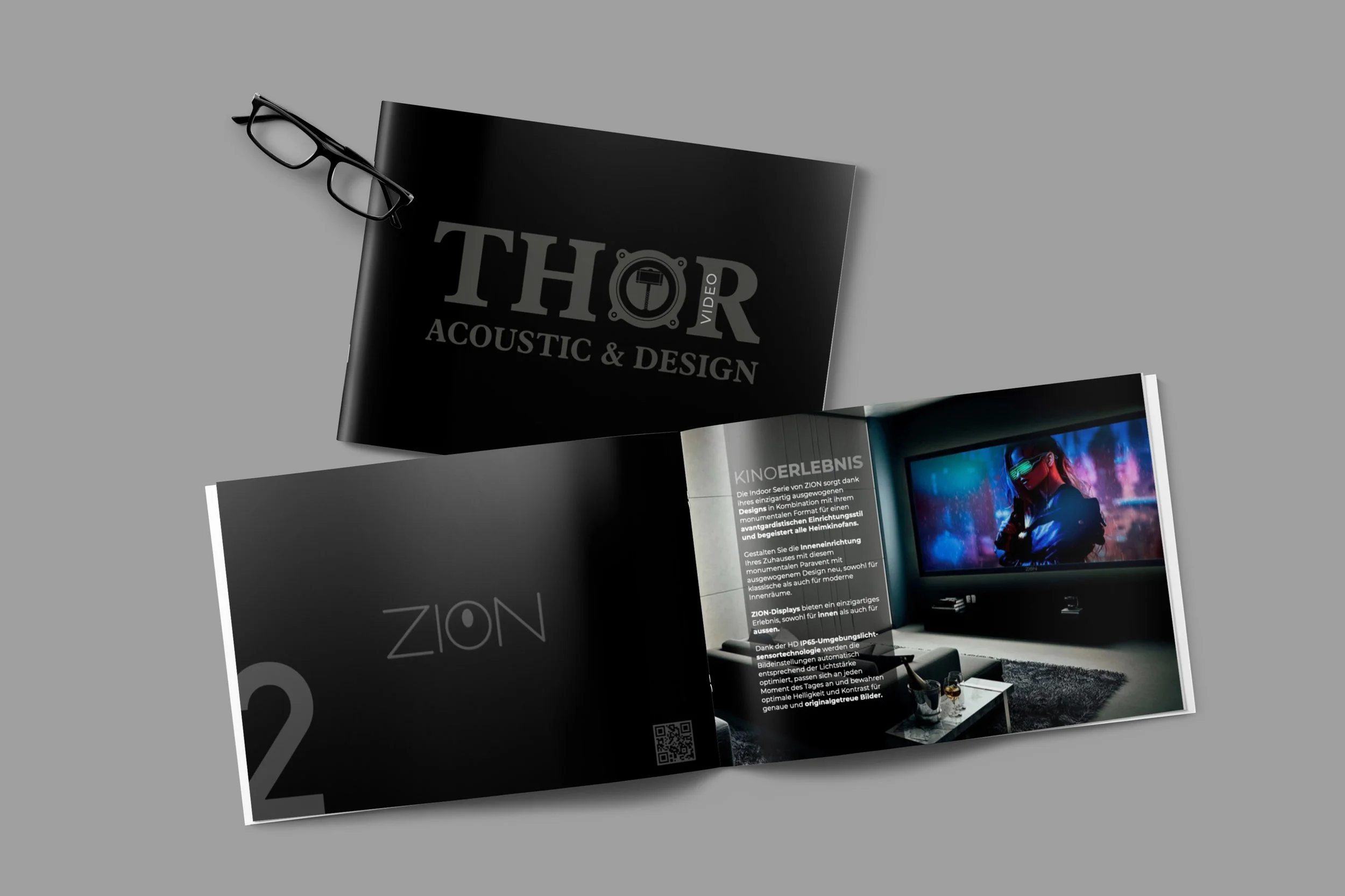 Offene Broschüre über Innenarchitektur und Design mit einem Bild eines Heimkino-Raums und Text auf der rechten Seite, schwarze Titelseite mit Logo, schwarze Lesebrille auf schwarzem Cover