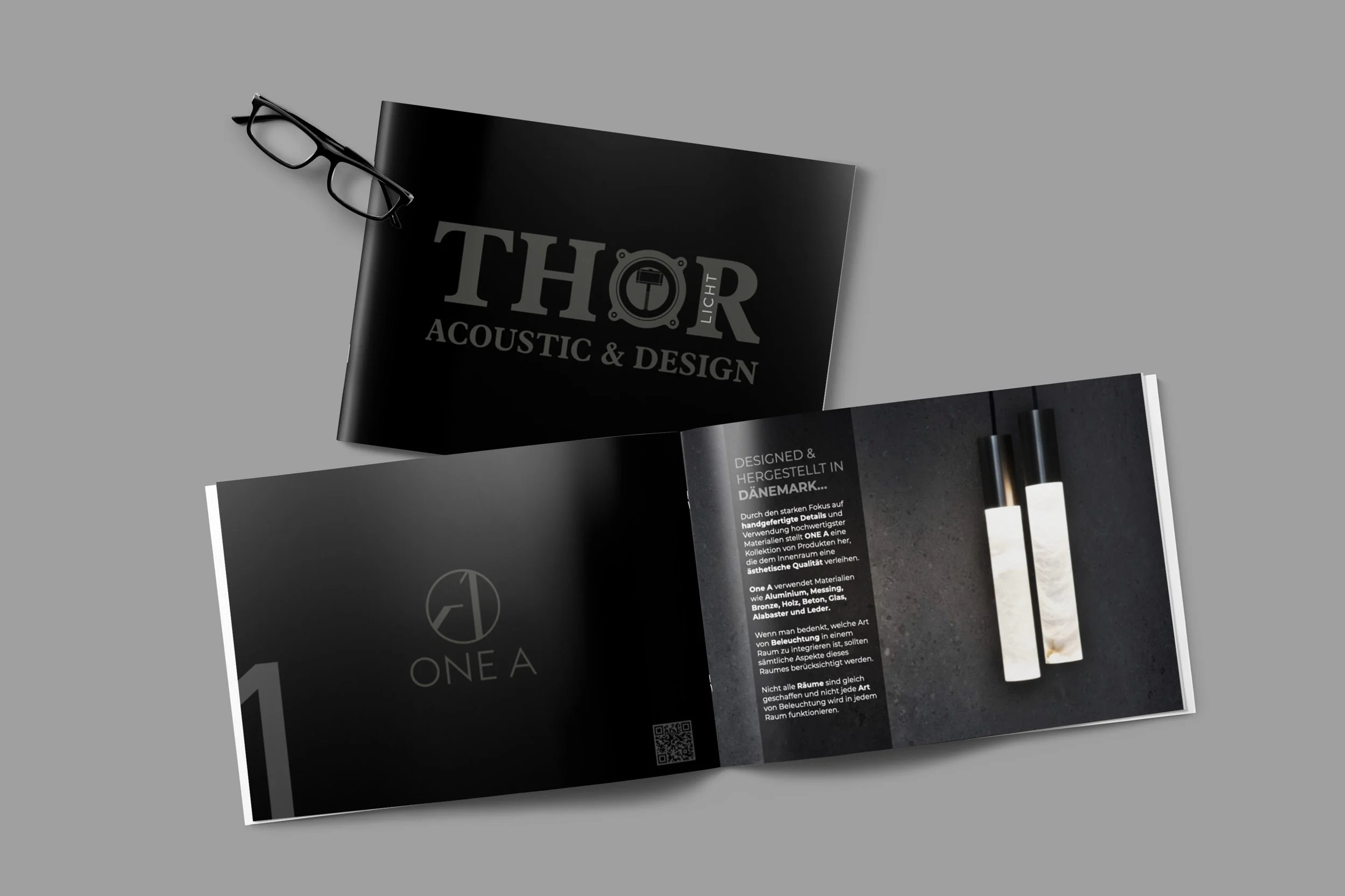 Offen geblättertes schwarzes Info-Buch, das das Logo 'ONE A' zeigt, neben einem schwarzen Magazin mit dem Titel 'THOR ACOUSTIC & DESIGN' und eine Lesebrille auf einem grauen Hintergrund.