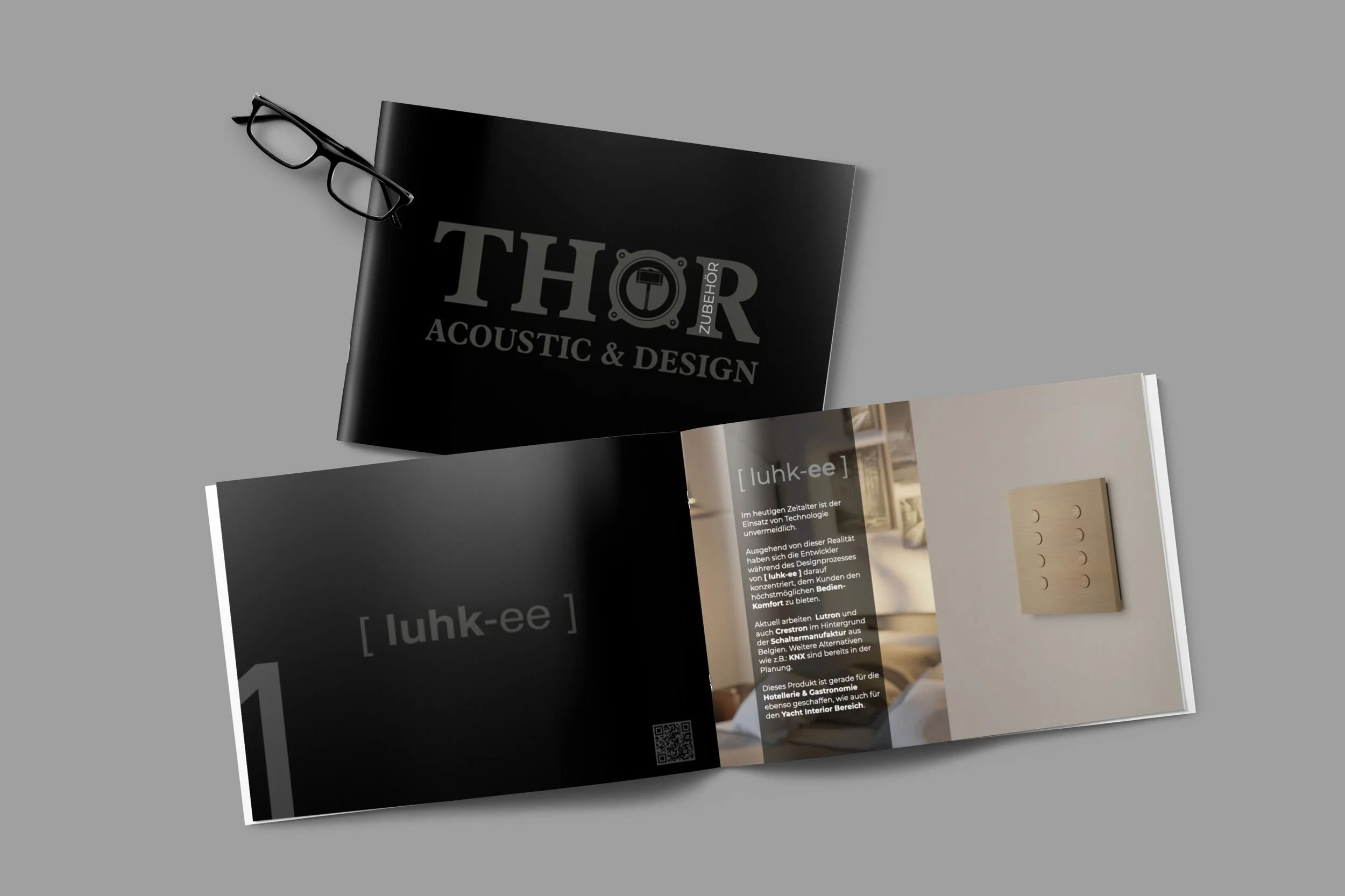Offene Broschüre für ein Produkt namens 'luhk-ee', mit deutschem Text und einem Bild eines Stromschalters auf der rechten Seite, neben einer schwarzen Broschüren mit 'THOR acoustic & design' auf einem grauen Hintergrund, daneben liegt eine schwarze Brille.