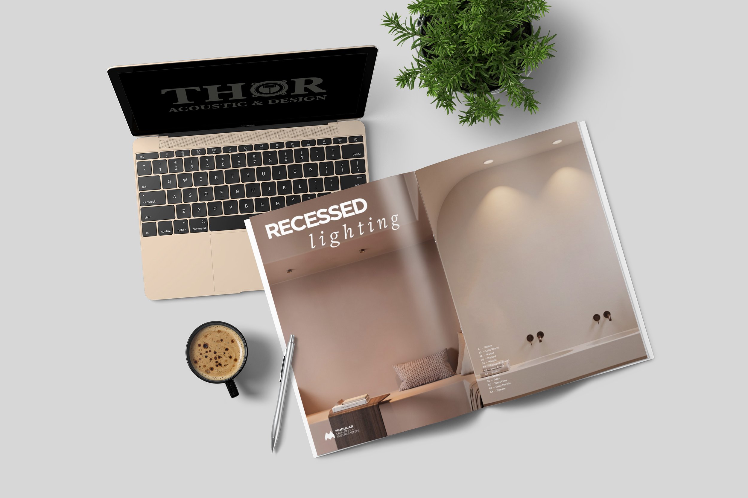 Arbeitsplatz mit Laptop, Kaffee, Stift, Magazin mit der Überschrift Recessed Lighting, und einer Zimmerpflanze auf hellem Tisch.