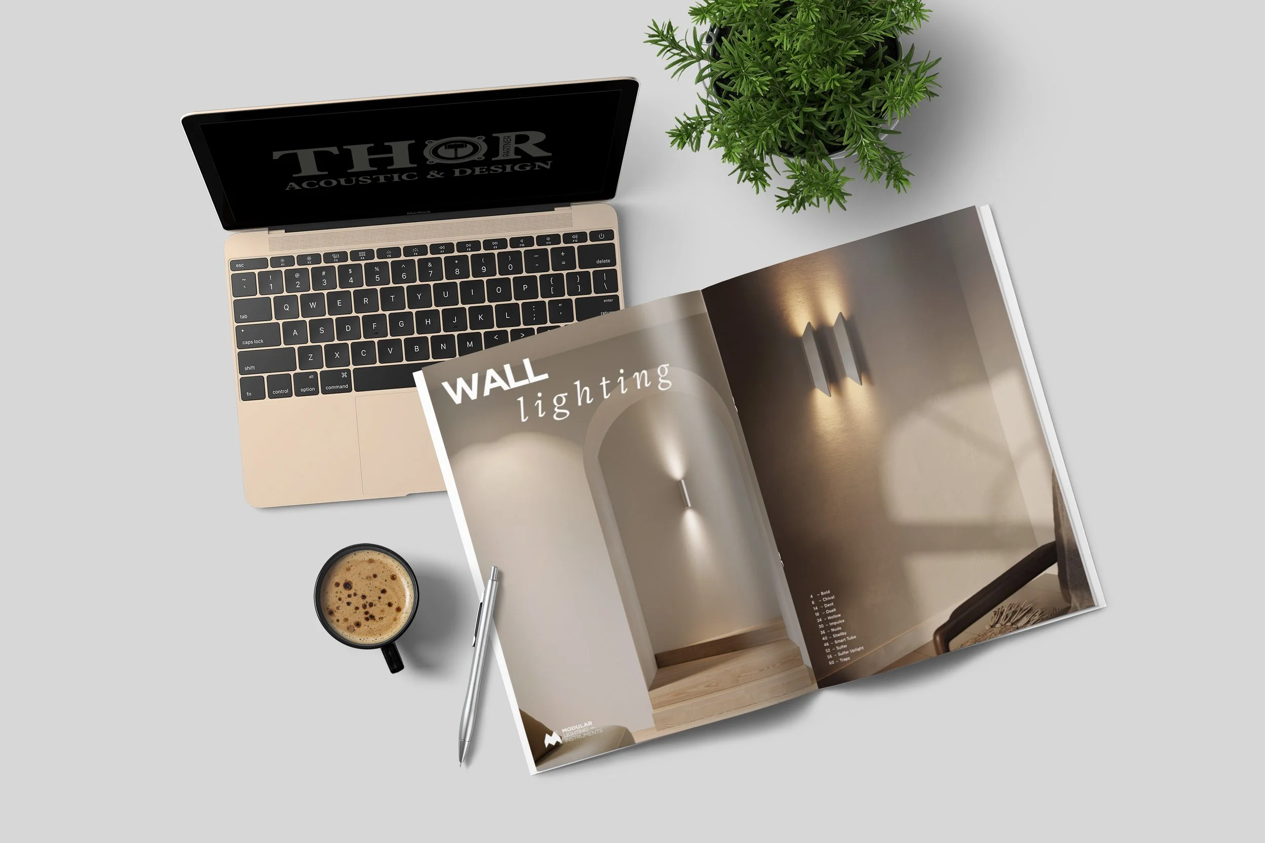 Arbeitsplatz mit einem offenen Katalog über Wandbeleuchtung, Laptop, Kaffeetasse, Kugelschreiber, Notizbuch und Zimmerpflanze auf einem hellgrauen Tisch.