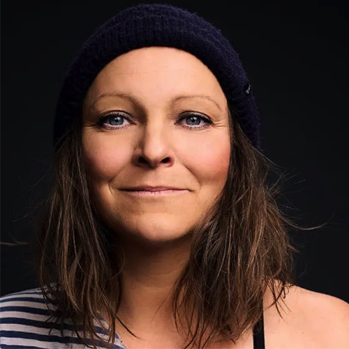 Nahaufnahme einer lächelnden Frau mit braunen Haaren, dunklem Beanie und gestreiftem Oberteil vor schwarzem Hintergrund.