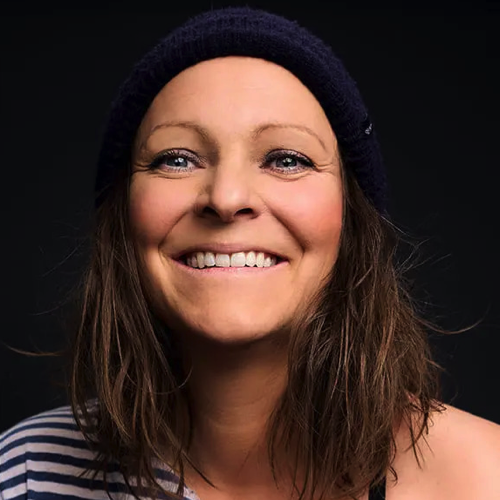 Portrait einer lachenden Frau mit braunen, schulterlangen Haaren, dunklem Beanie und gestreiftem Oberteil vor schwarzem Hintergrund.