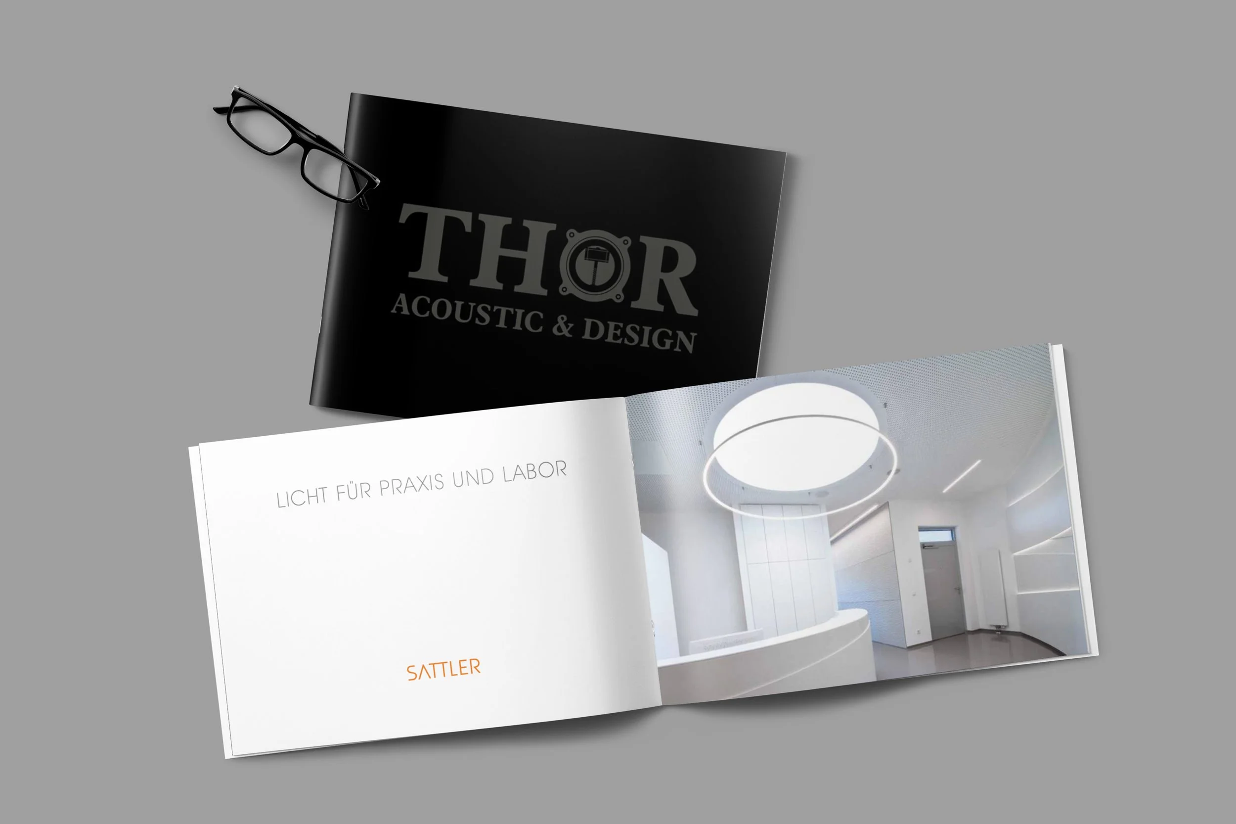 Aufgeschlagenes modern gestaltetes Design-Buch mit Innenraum-Ansicht, daneben schwarzes Buch oder Katalog mit Logo "THOR ACOUSTIC & DESIGN" und eine Brille