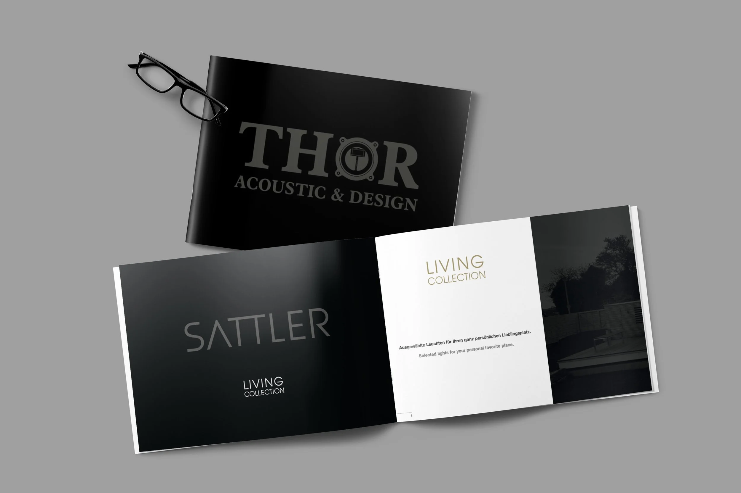 Offene Broschüre für die Sattler Living Collection mit dunklen und weißen Seiten, schwarzen Rahmen, und eine schwarze Brille liegt oben auf der Broschüre.
