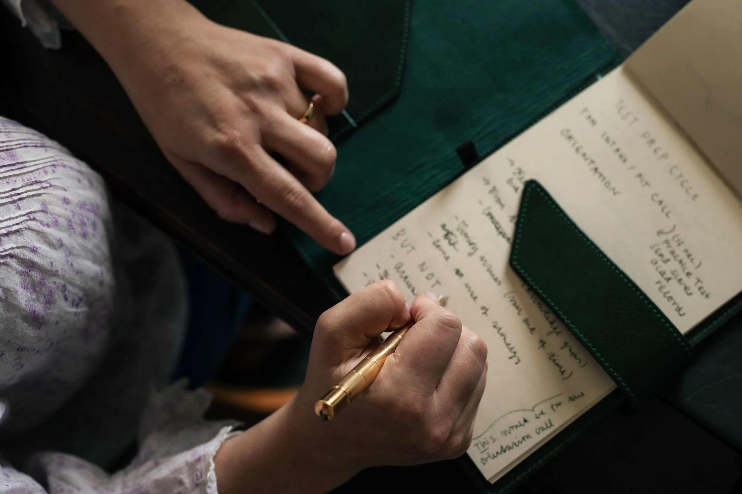 A woman's hand holding a gold pen writing in a notebook, next to a dark gray leather pen holder, on a table. Part of her arm and a corner of a purple patterned blouse are visible.