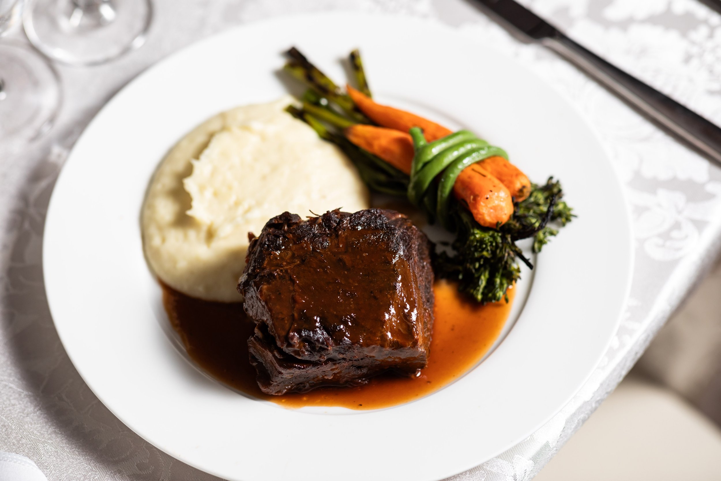A plated meal of steak with gravy, mashed potatoes, and roasted vegetables including carrots and broccolini on a white dish.