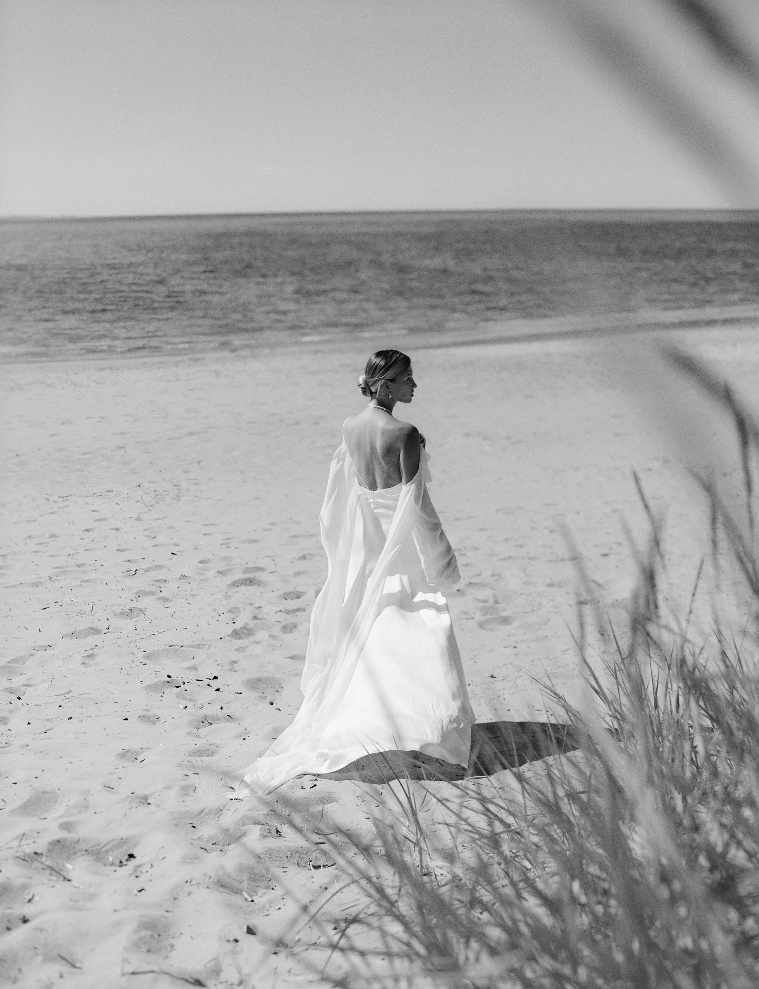 A woman in a wedding dress walking on a sandy beach, viewed from the side amidst some beach grass, with the ocean in the background.