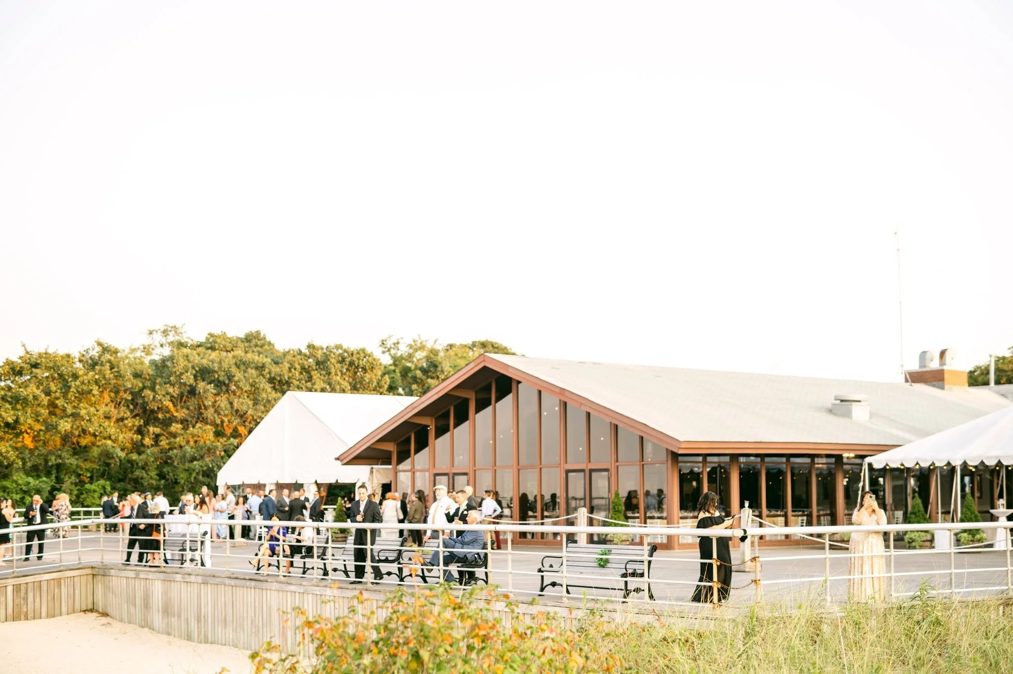 A gathering of people outside a large modern building with wooden accents, surrounded by trees, on a sunny day.